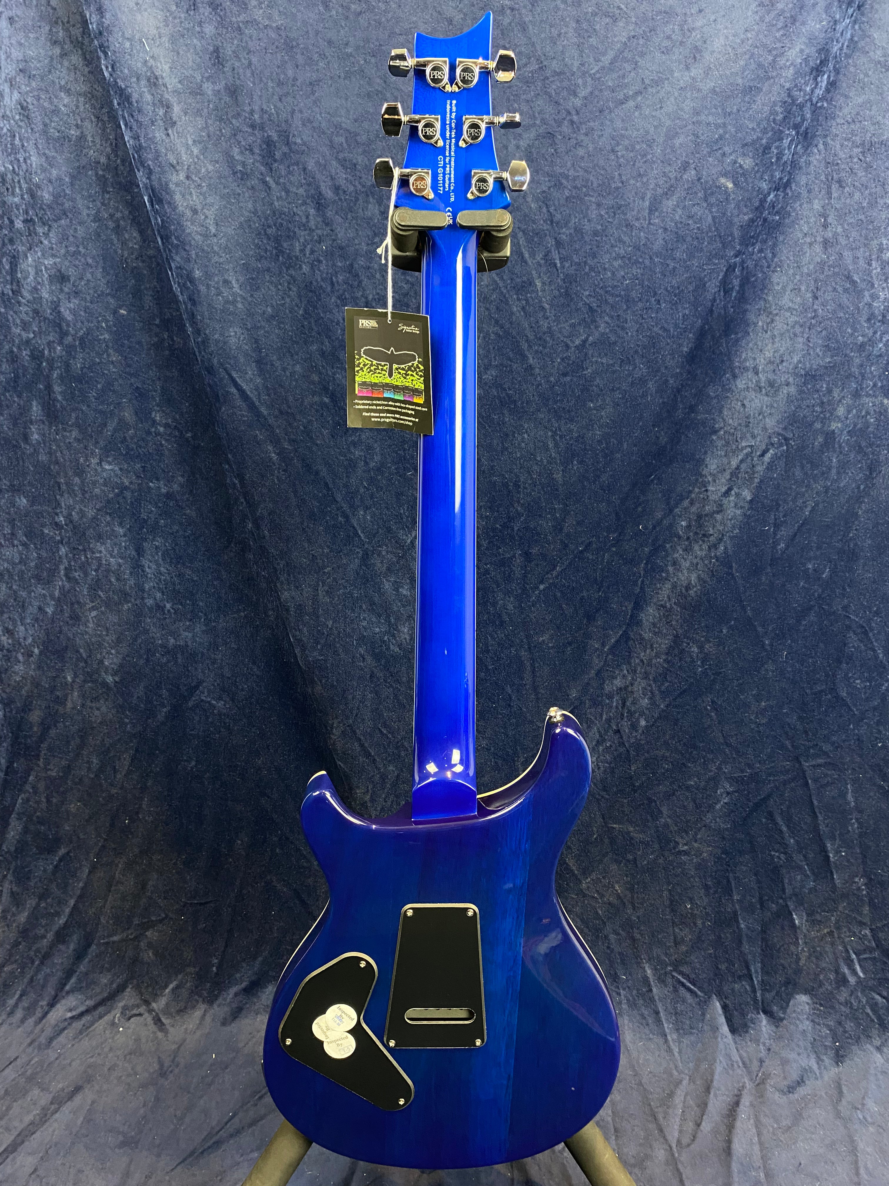 Bright blue electric guitar shown from the back, with chrome tuning pegs, two backplates, and a tag hanging from the headstock.
