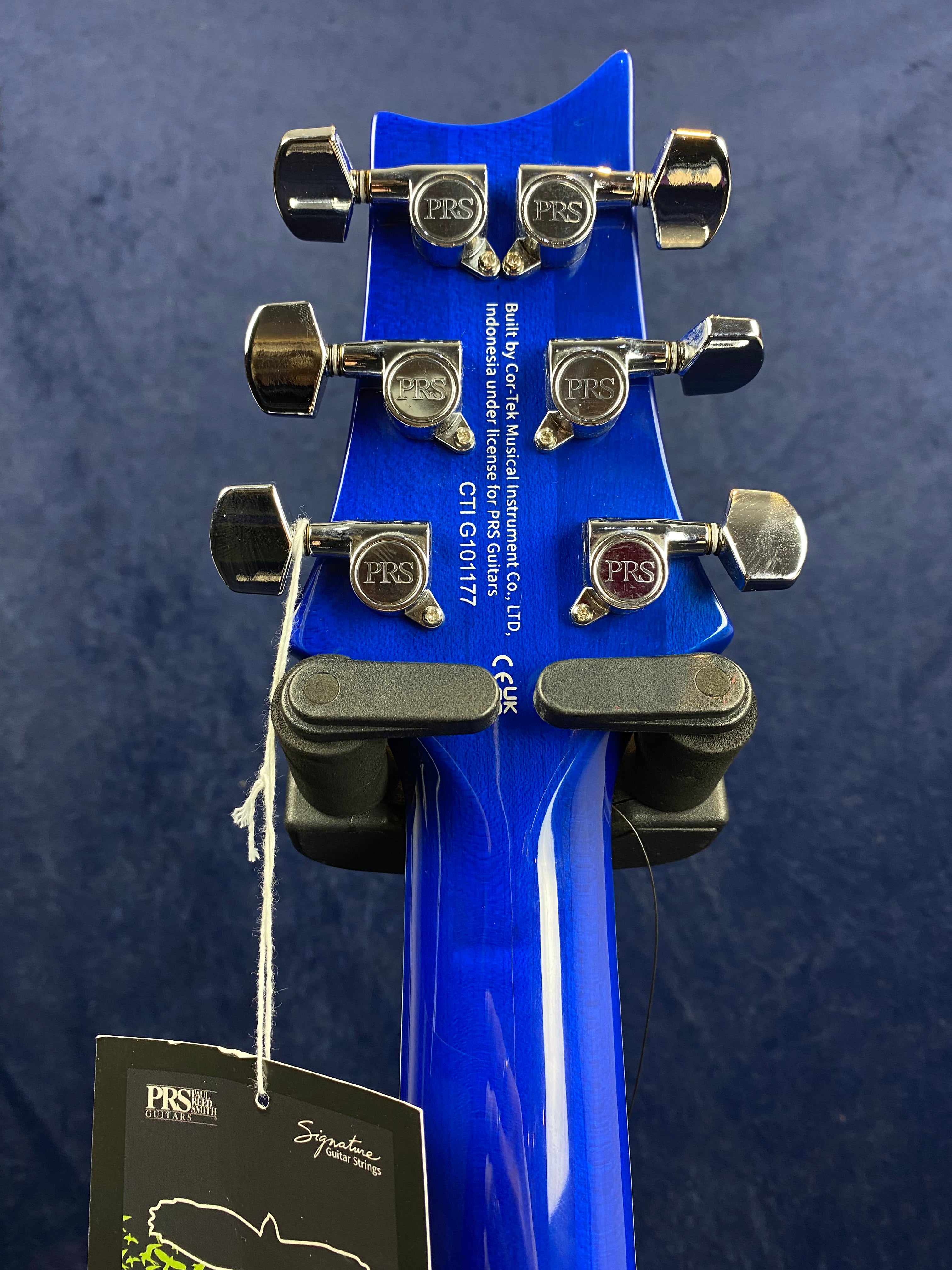 Blue electric guitar headstock with six chrome tuning pegs labeled PRS, product tag attached below, white serial number and text along the back.
