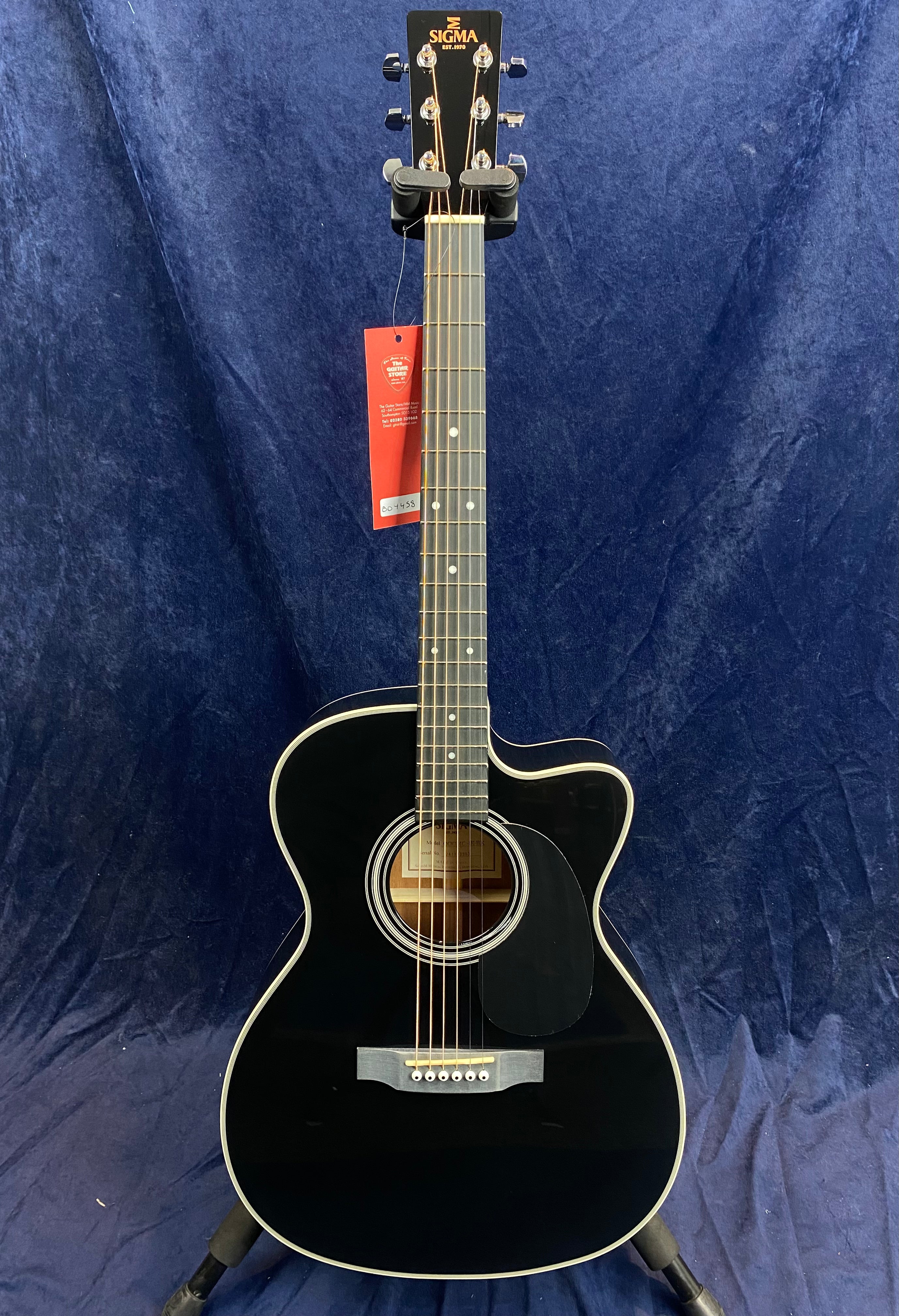 Black acoustic guitar with white trim, cutaway body, six strings, round sound hole, and a red tag hanging from the neck.