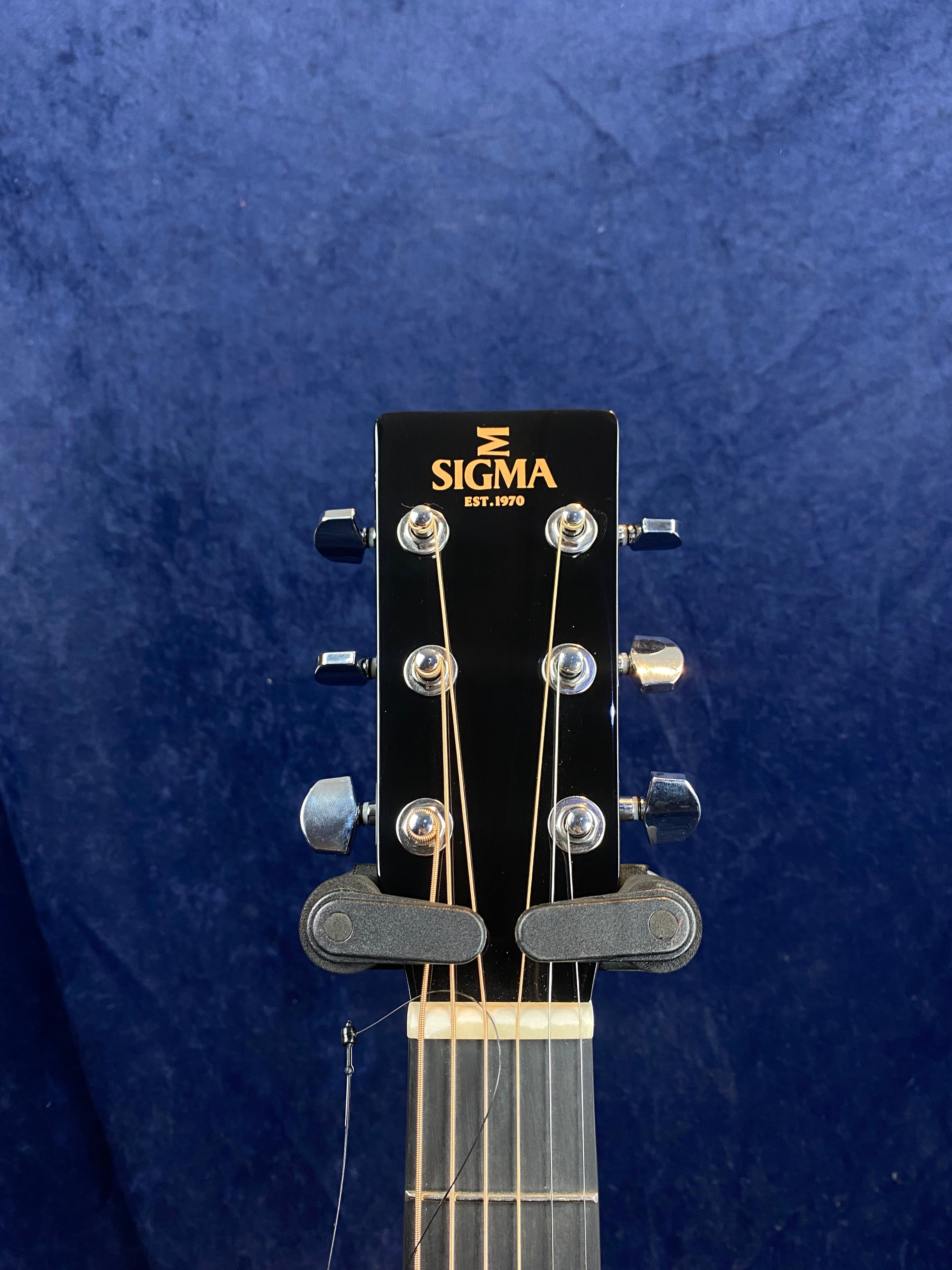Headstock of a Sigma acoustic guitar with tuning pegs, strings, and the Sigma logo on a dark background.
