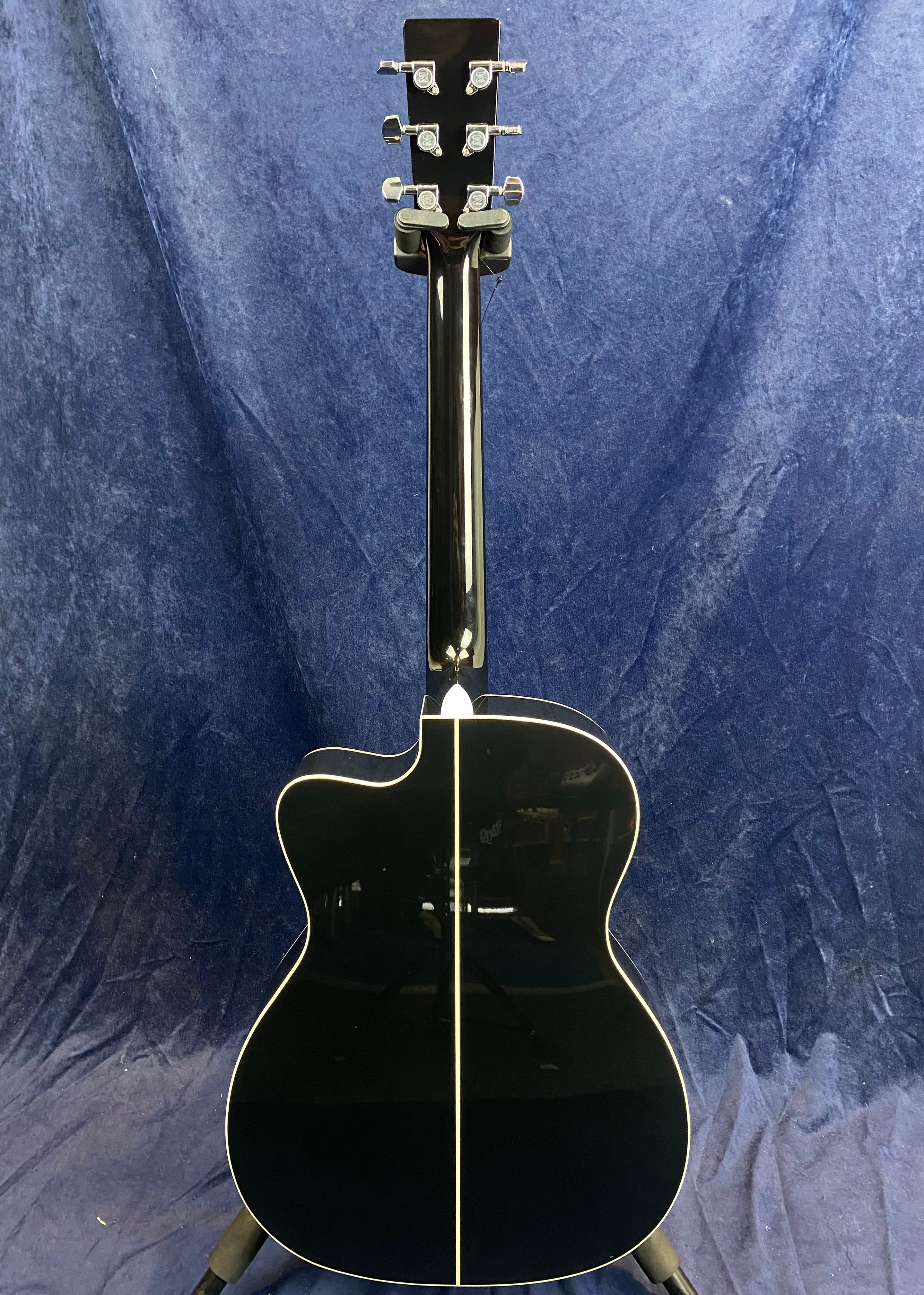 Black acoustic guitar shown from the back, with a white center stripe, cream edge binding, and metal tuning pegs against a dark blue background.