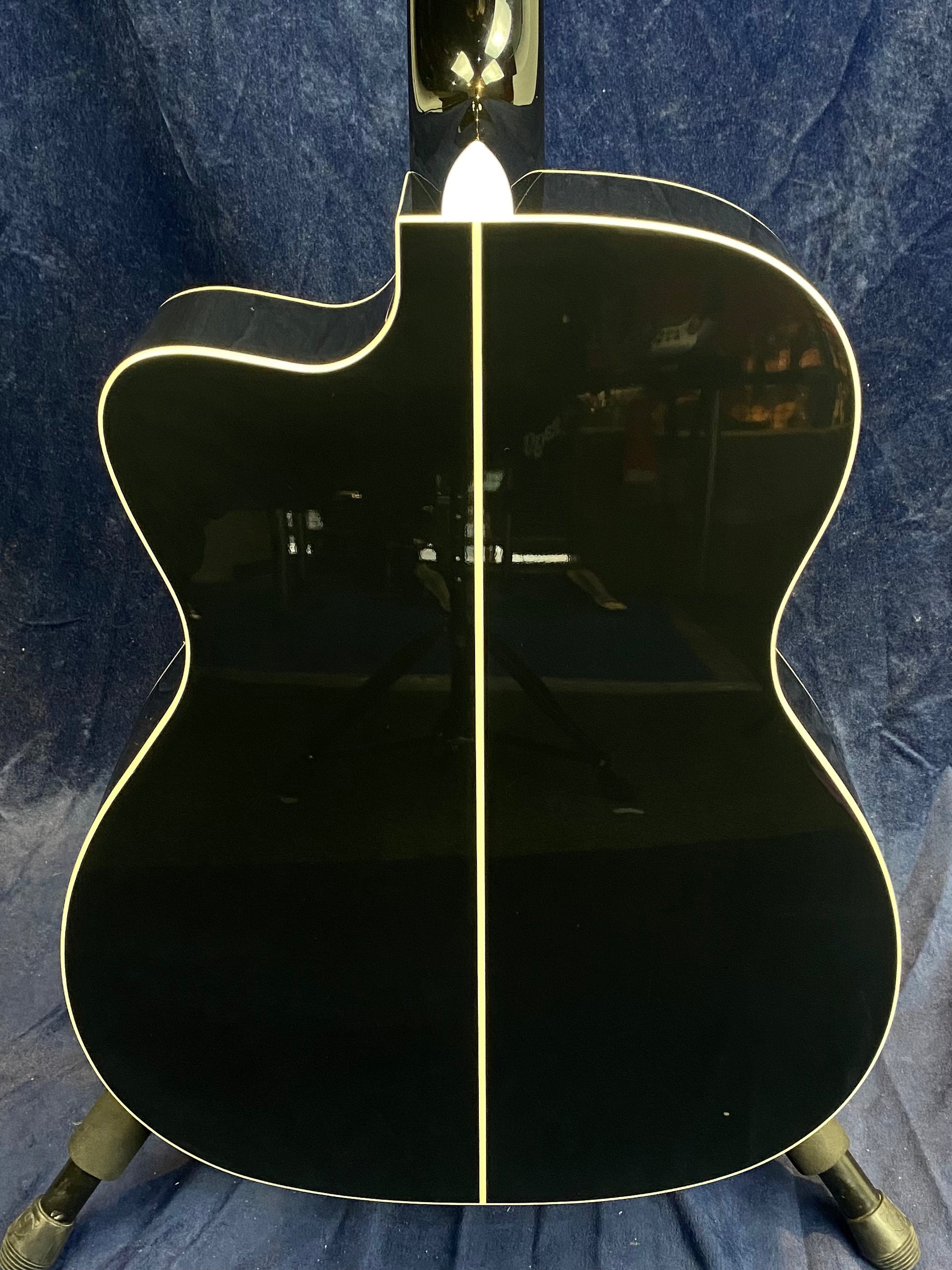 The back of a glossy black acoustic guitar with cream binding and a vertical cream stripe down the center, set against a dark blue fabric background.