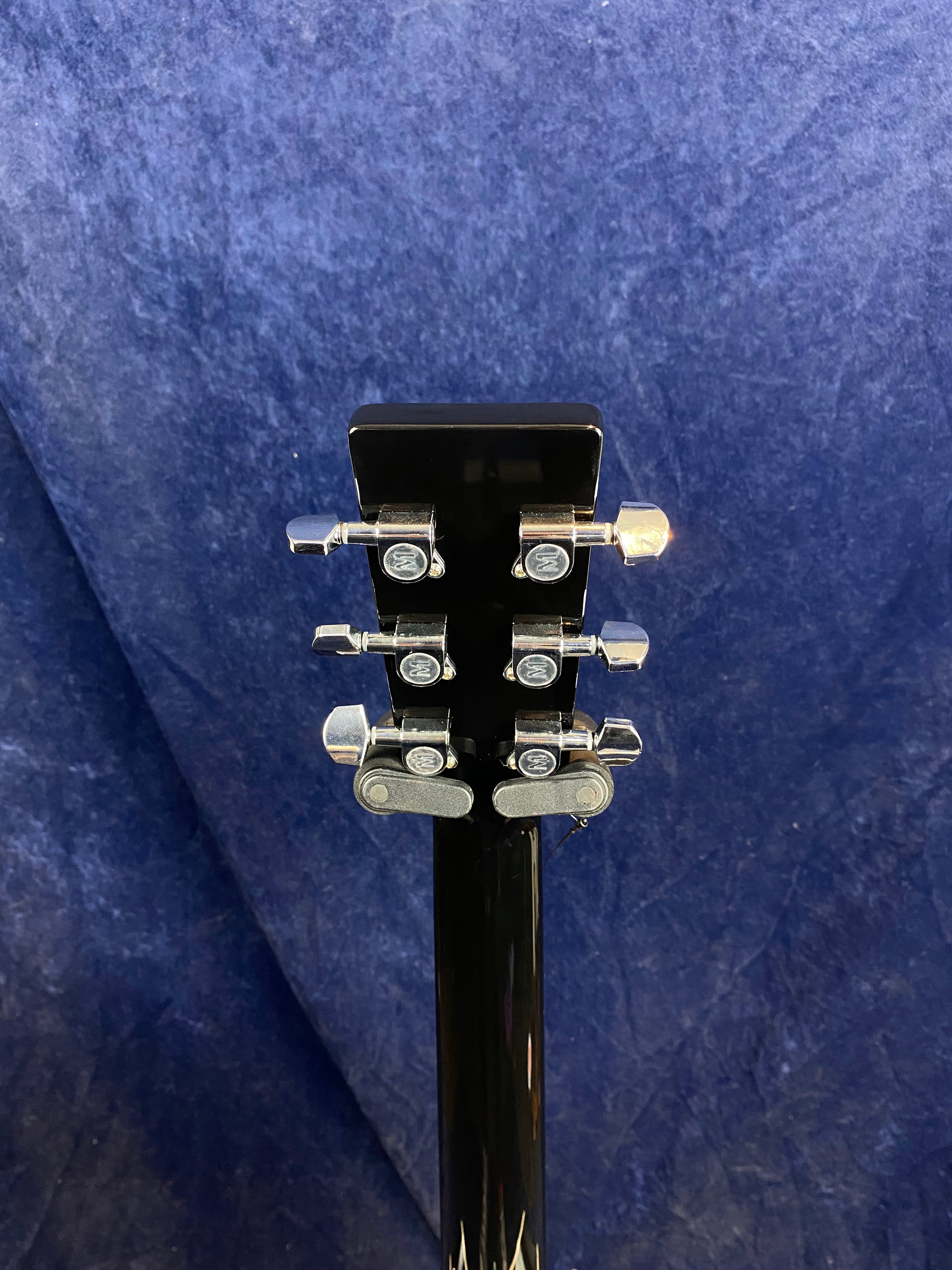 Close-up of the back of an acoustic guitar headstock showing six chrome tuning pegs against a dark blue textured background.