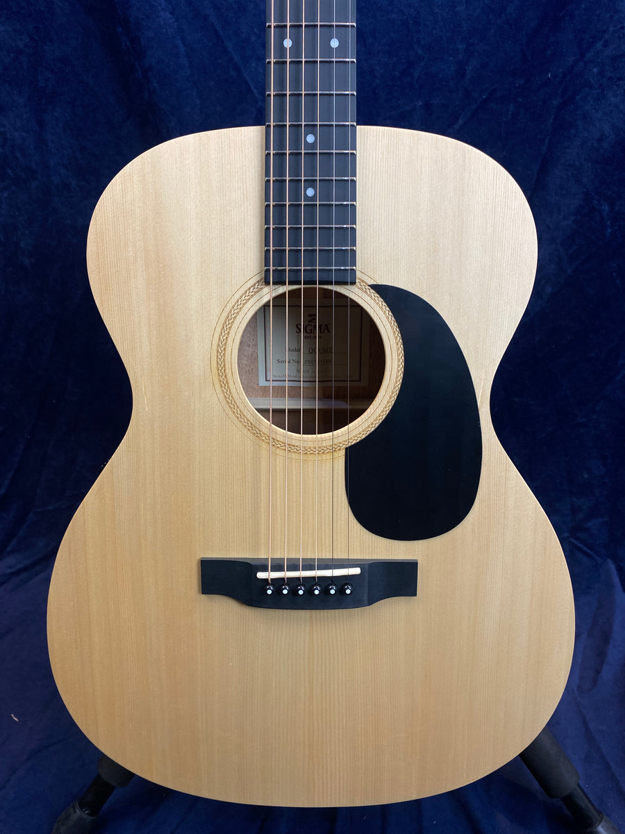 Light wood acoustic guitar with six strings, a black pickguard, and dot inlays on a dark fretboard, shown against a dark blue background.