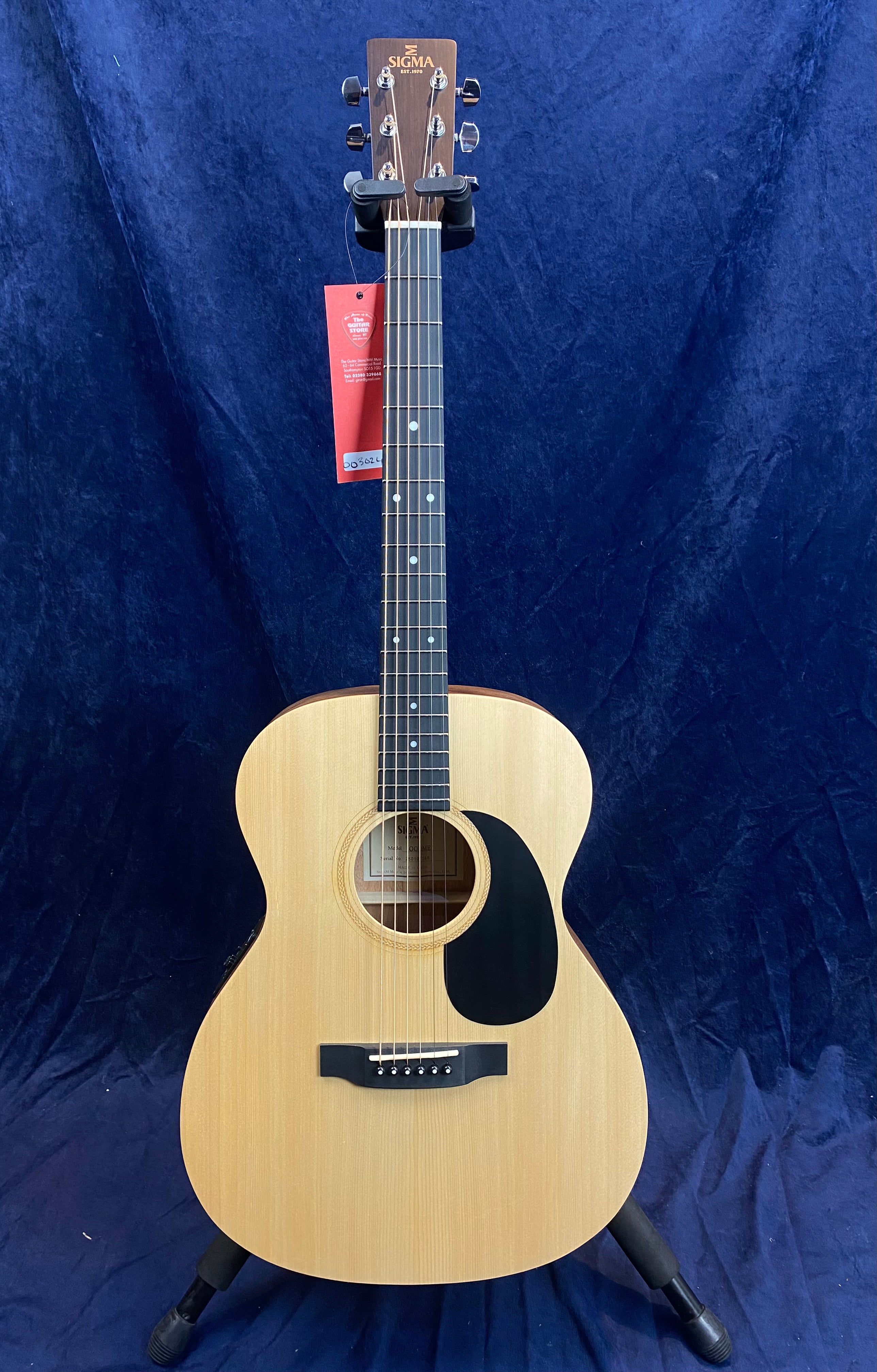 Light wood acoustic guitar with a black pickguard, six strings, round sound hole, and a Sigma brand logo on the headstock, displayed on a stand.