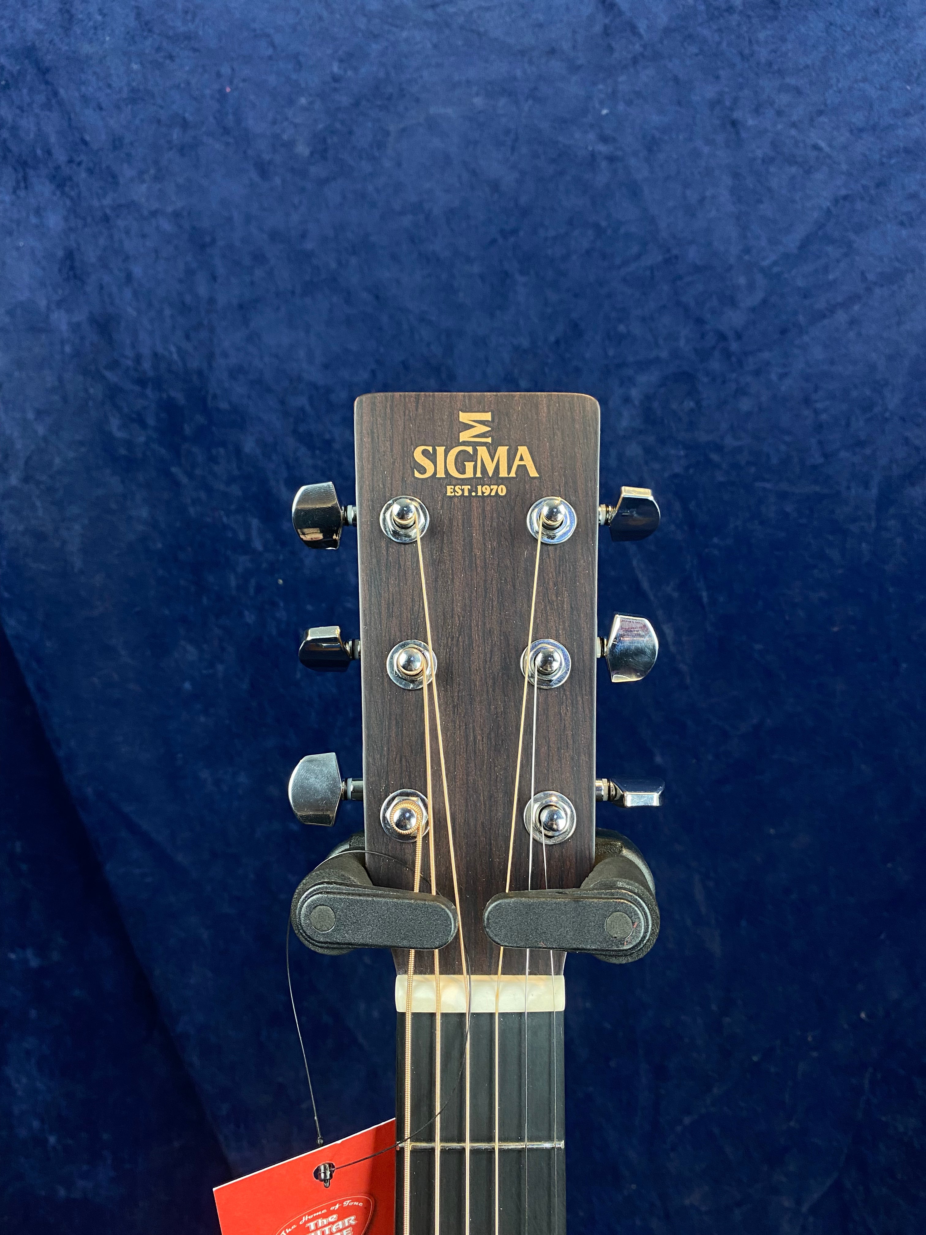 The headstock of a Sigma acoustic guitar with six tuning pegs, "Sigma Est. 1970" logo, and a string tag hanging below.