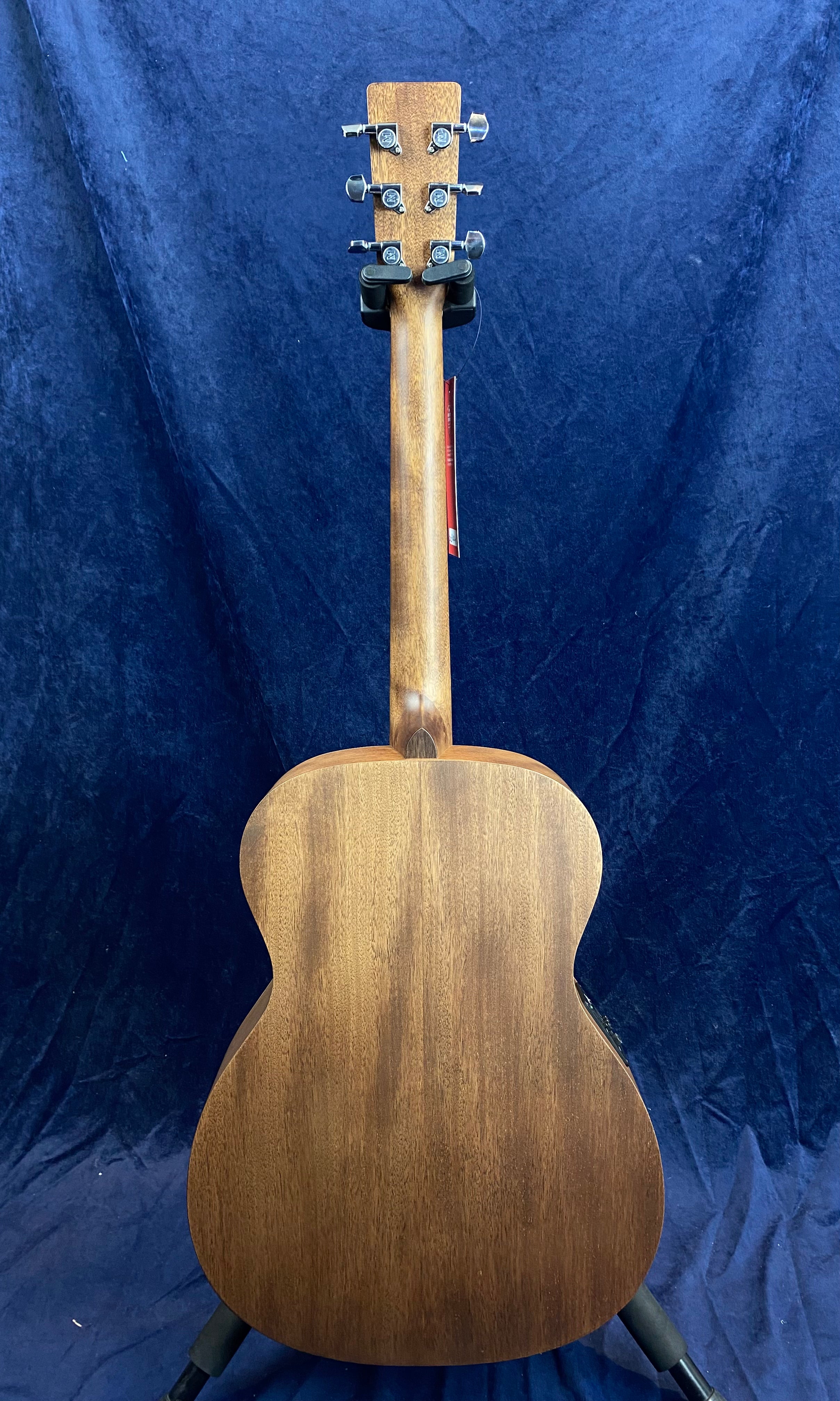 Acoustic guitar seen from the back, showing smooth brown wood grain, six silver tuning pegs, and resting on a stand against a dark blue background.