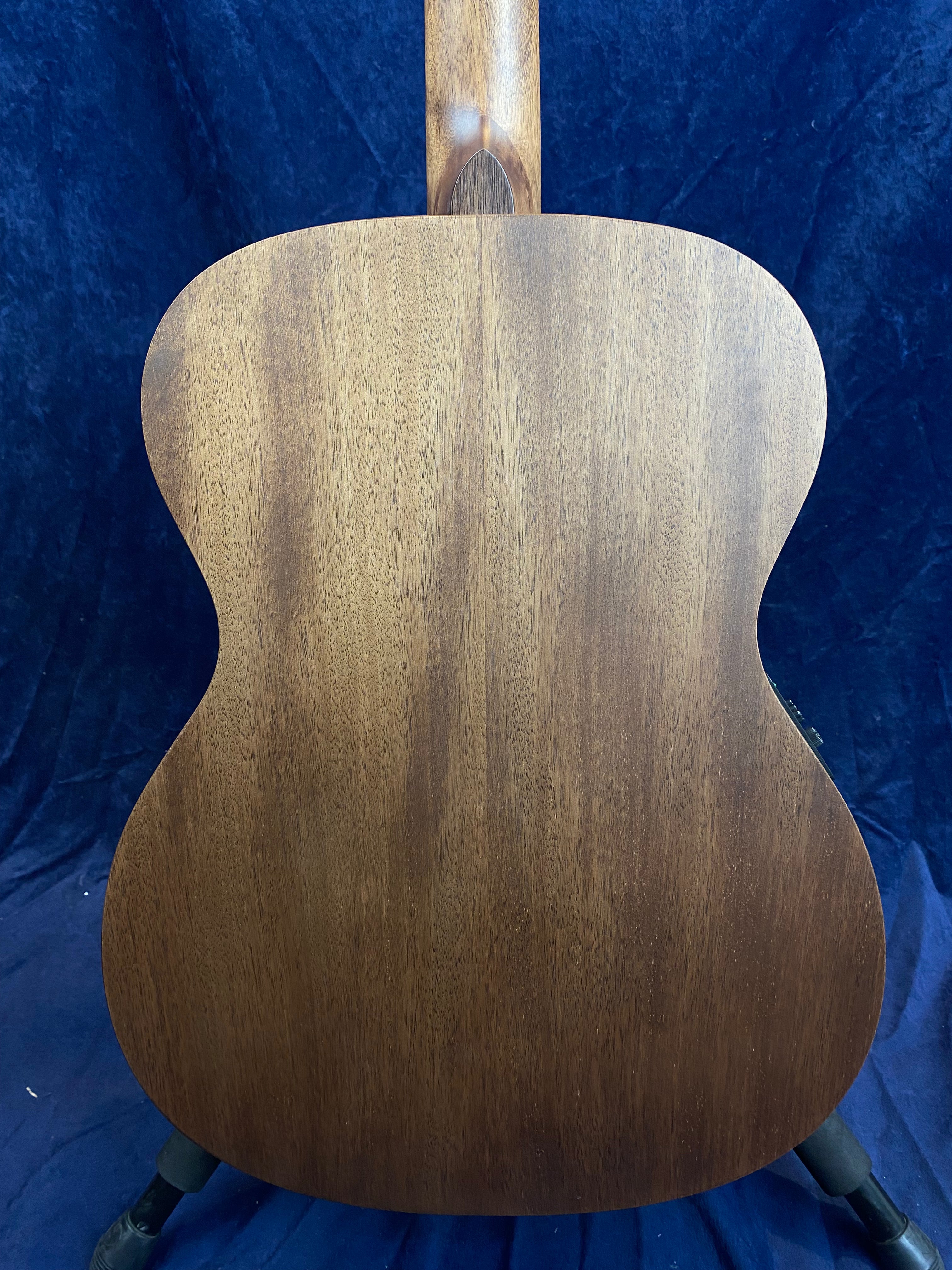 Acoustic guitar back made of smooth, medium-brown wood with subtle grain patterns, shown upright against a dark blue background.