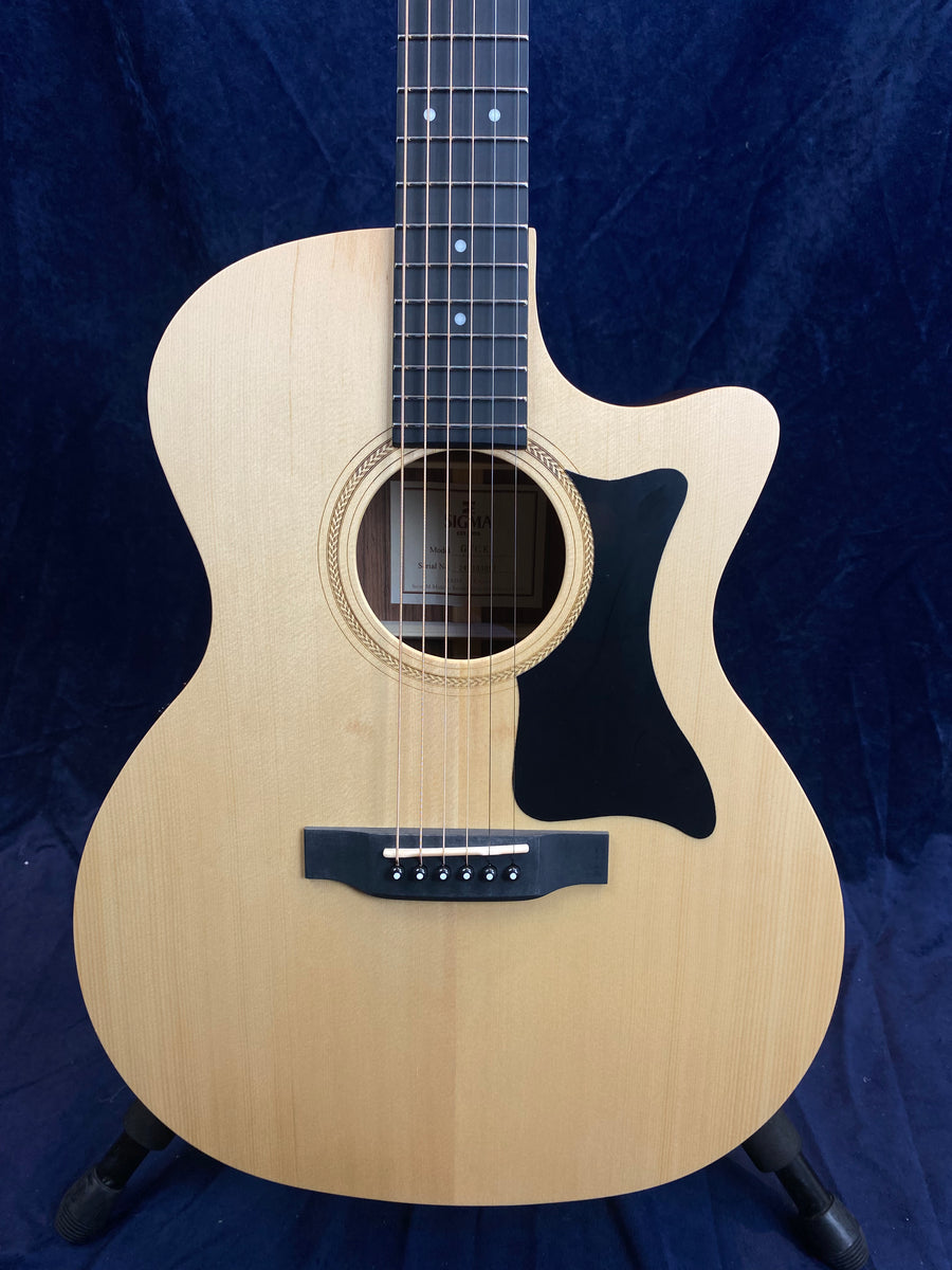 Light wood acoustic guitar with a black pickguard and cutaway design, six steel strings, dark bridge, and decorative soundhole rosette.
