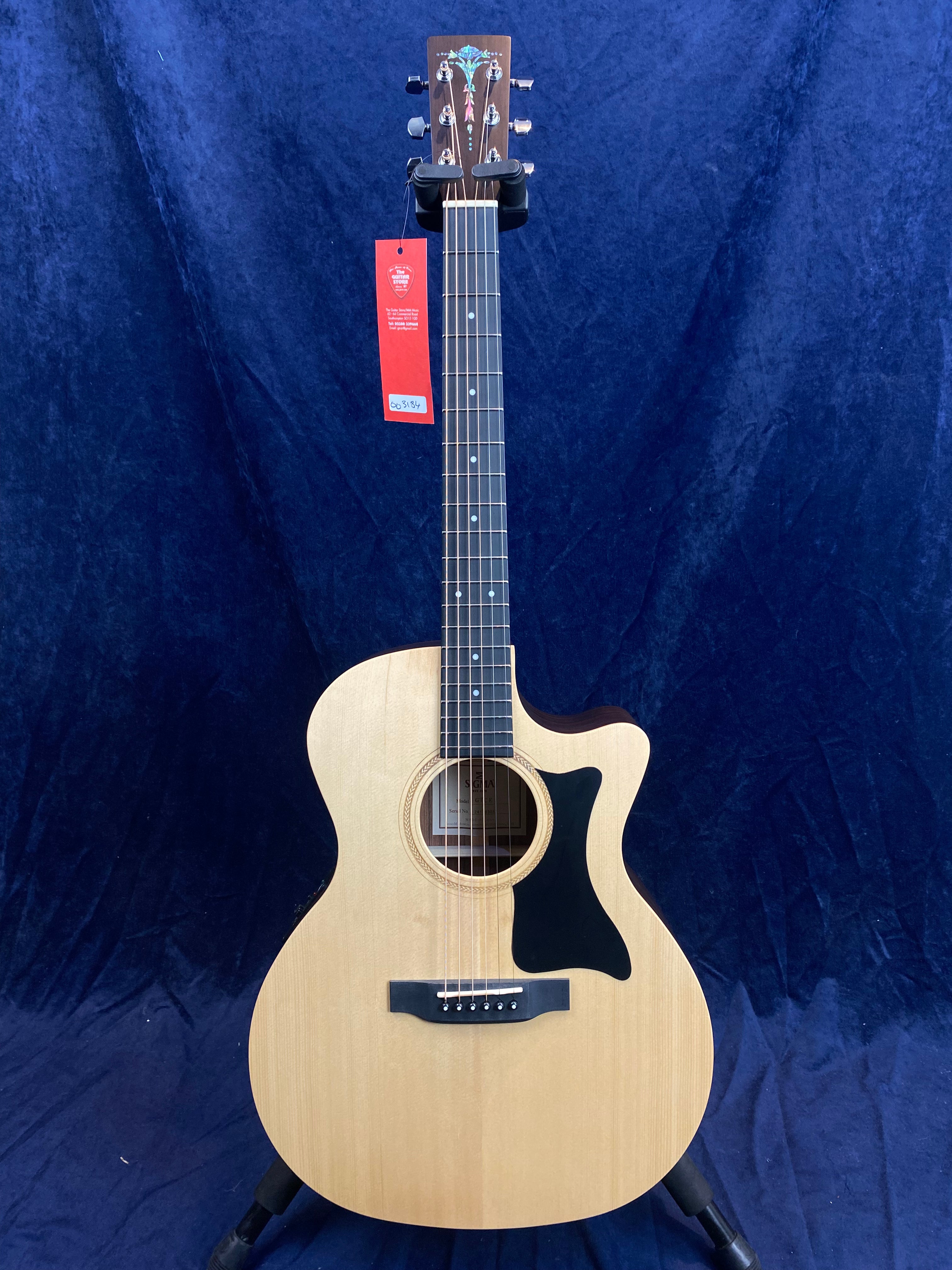 Natural wood acoustic guitar with a black pickguard, intricate headstock inlay, and a red tag hanging from the tuning pegs.