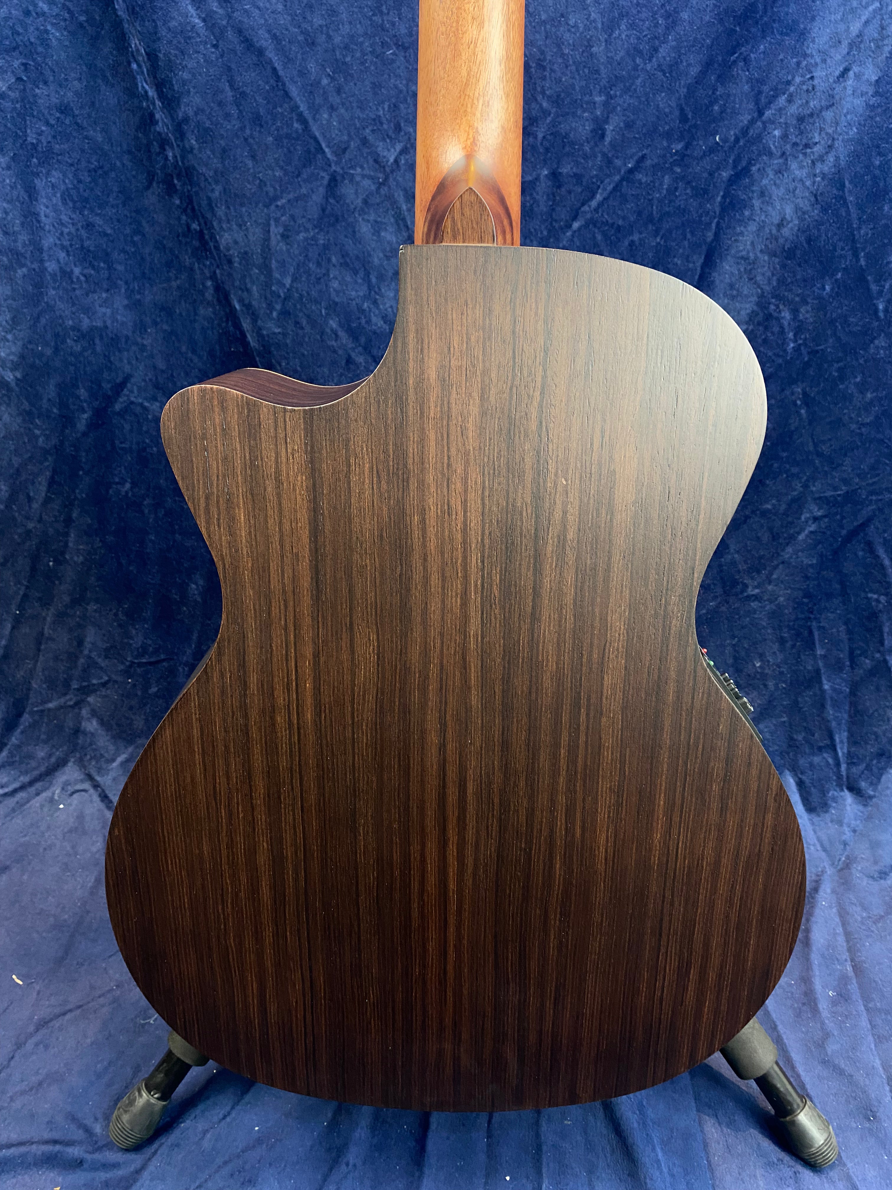 Dark wood acoustic guitar back with a smooth finish, gentle wood grain pattern, and rounded cutaway near the neck, shown upright on a stand.