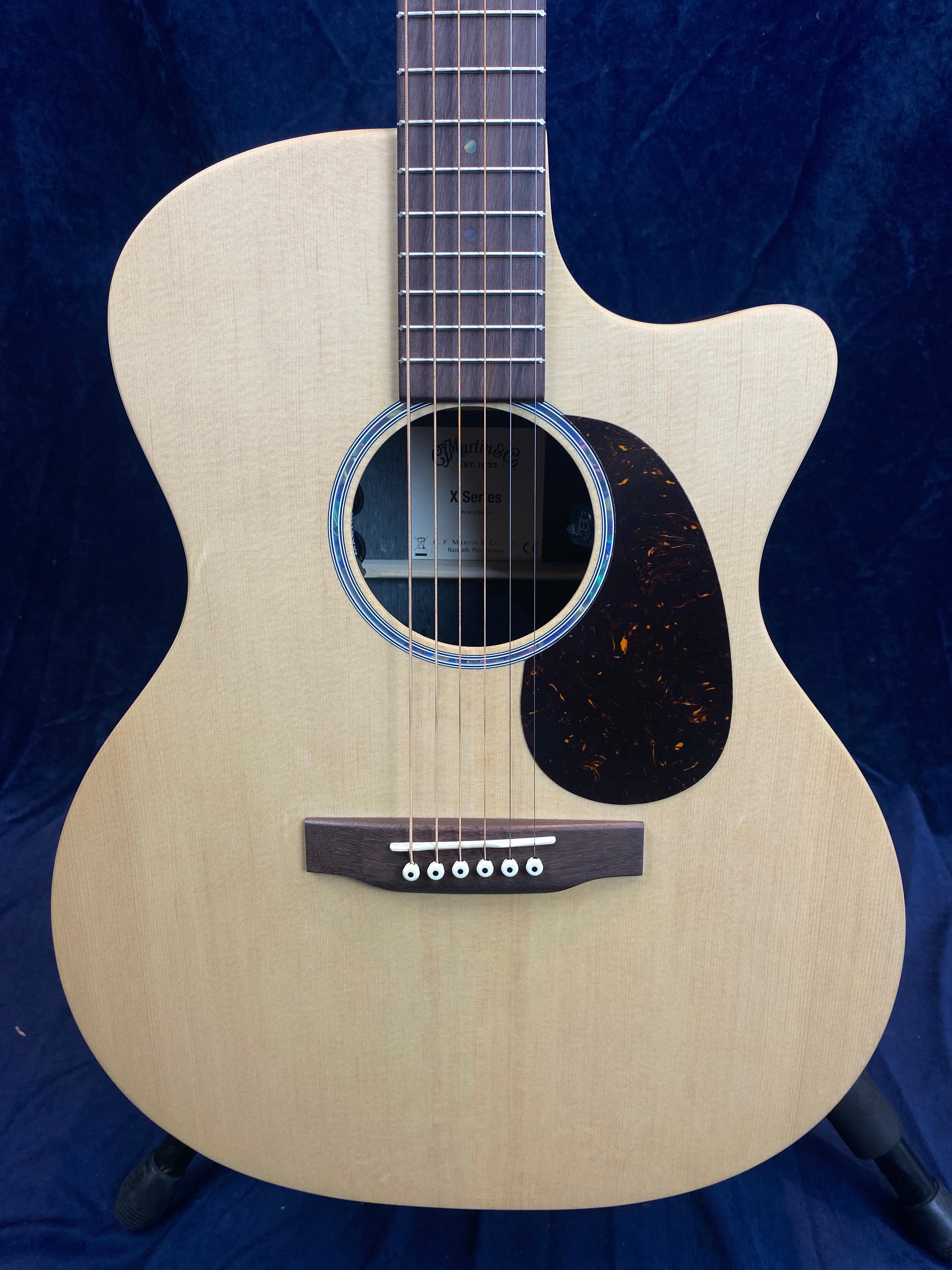 Light wood acoustic guitar with a cutaway body, dark pickguard, six strings, and a round sound hole with visible internal label.