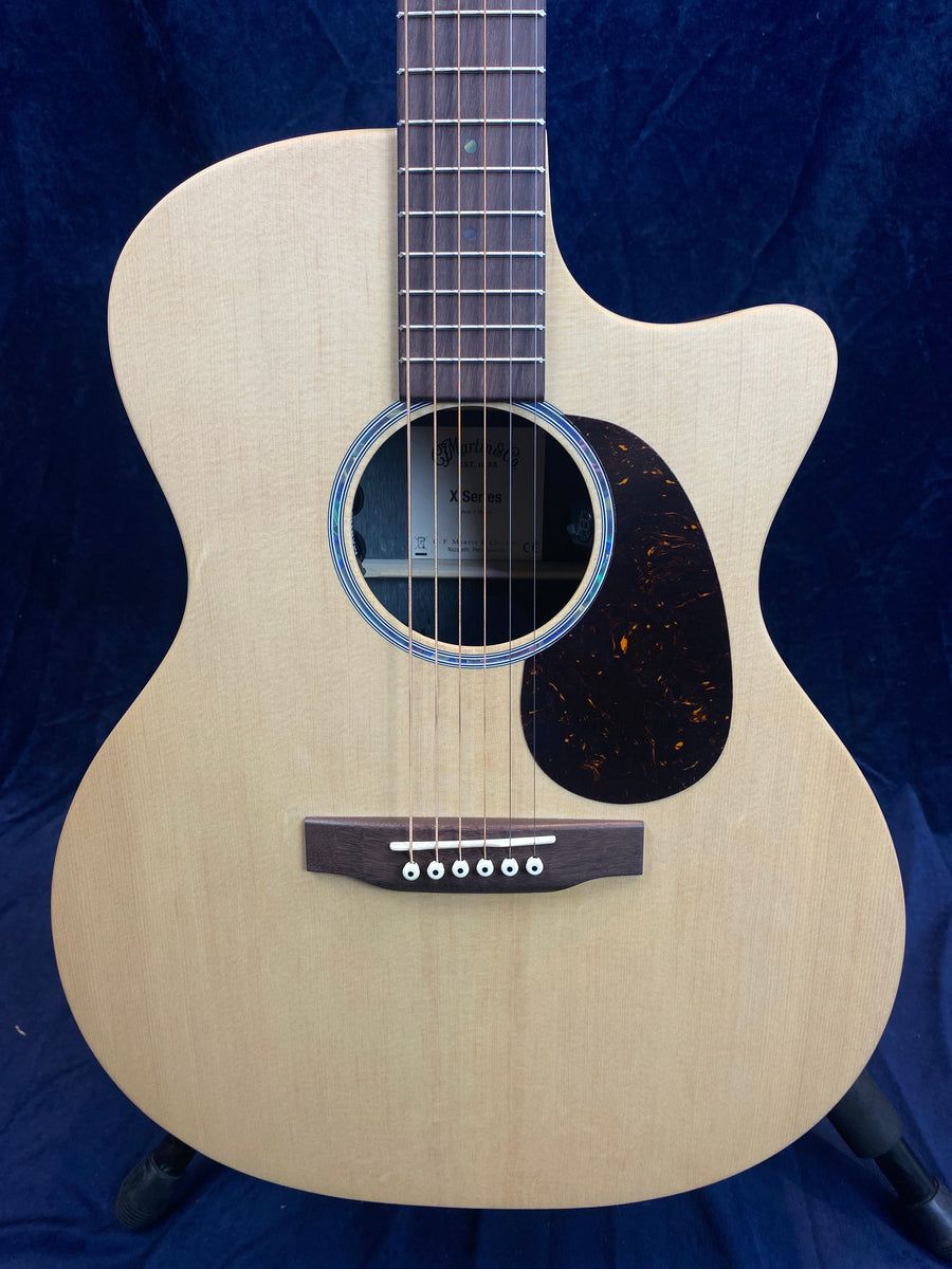 Light wood acoustic guitar with a cutaway body, dark pickguard, six strings, and a round sound hole with visible internal label.