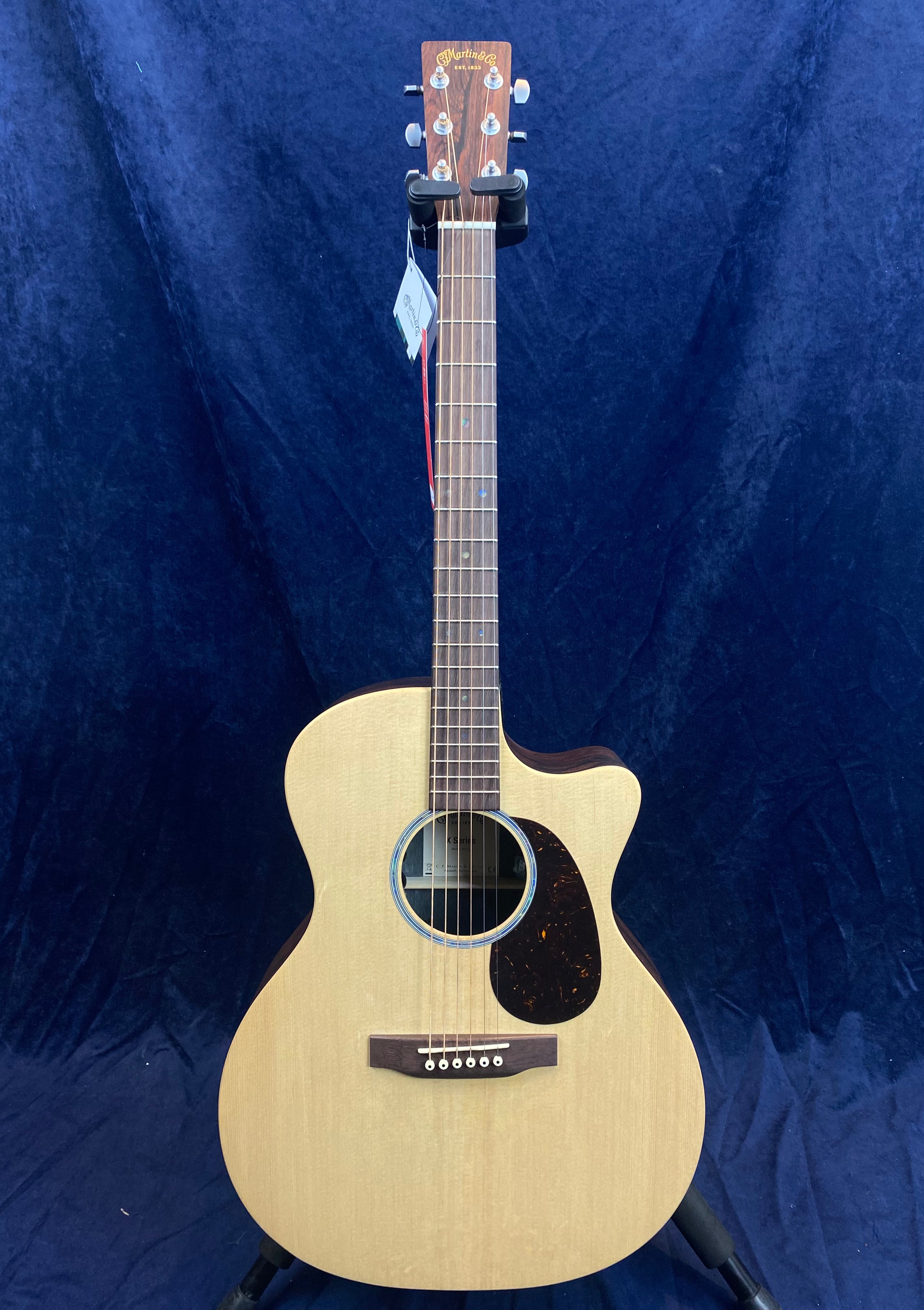 Natural wood acoustic guitar with a single cutaway, dark pickguard, six strings, and a tag hanging from the headstock.