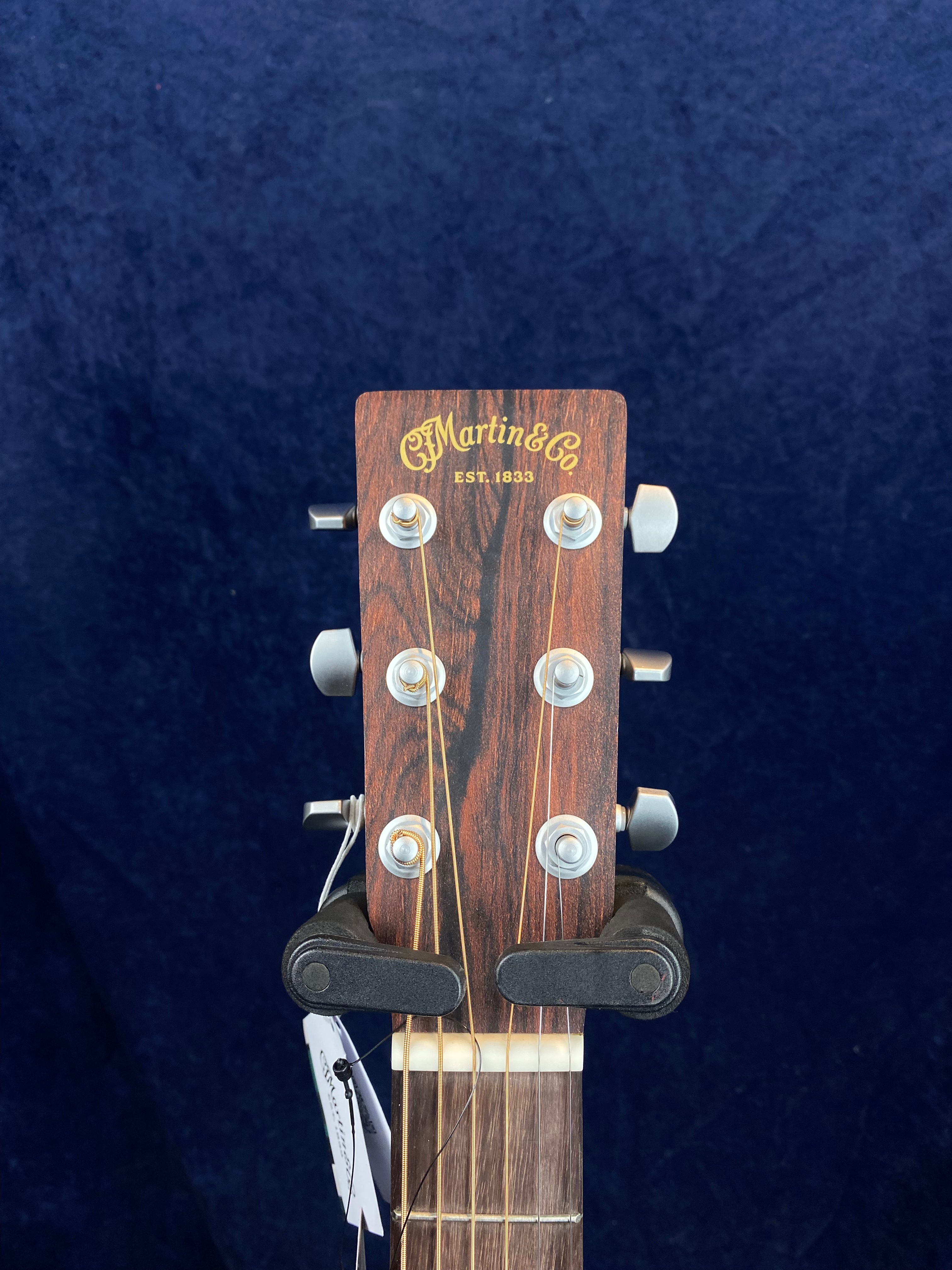 The headstock of a Martin & Co acoustic guitar with six tuning pegs, wooden finish, and gold logo text reading "Est. 1833" against a dark background.