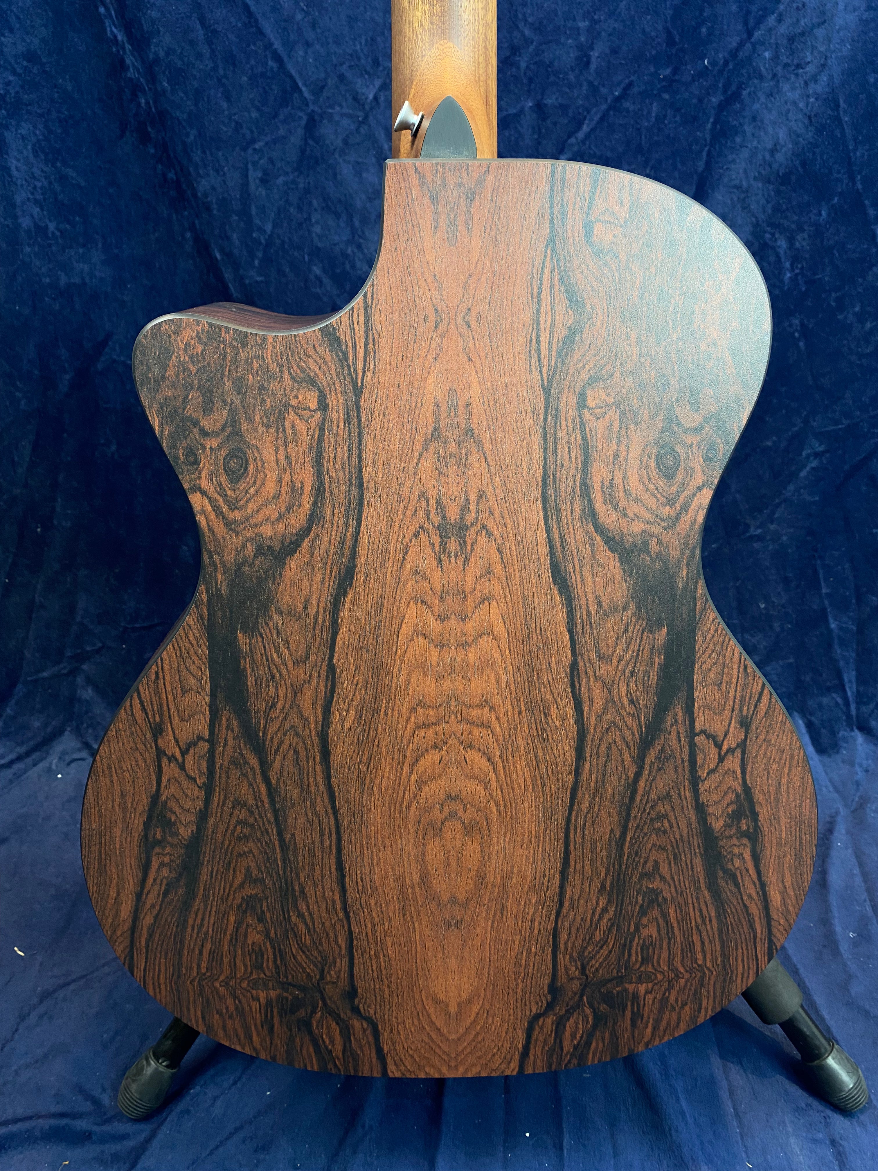 The back of an acoustic guitar shows rich brown wood with dramatic, dark, symmetrical grain patterns, standing against a dark blue background.