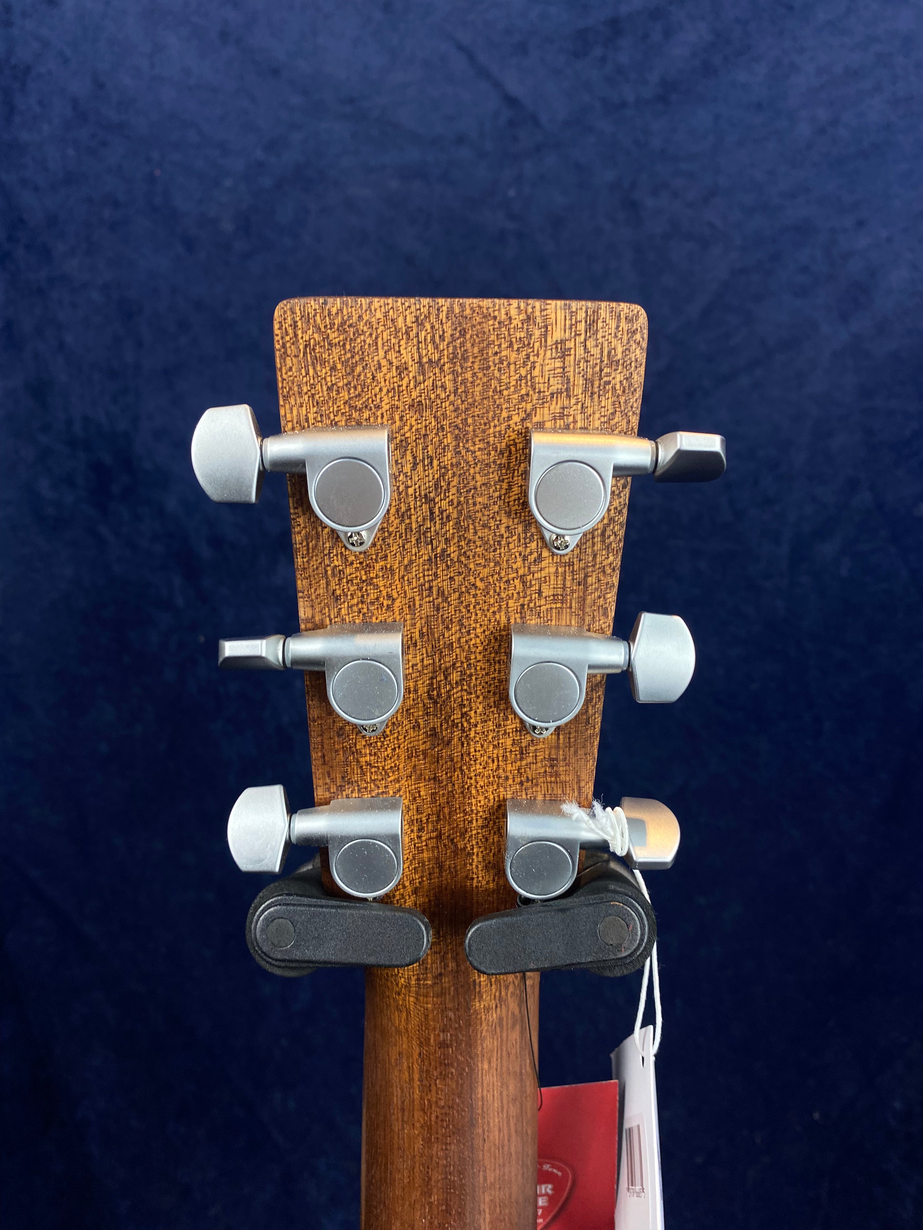 Rear view of an acoustic guitar’s headstock with six silver tuning pegs, a wood grain finish, and tags hanging from the neck.