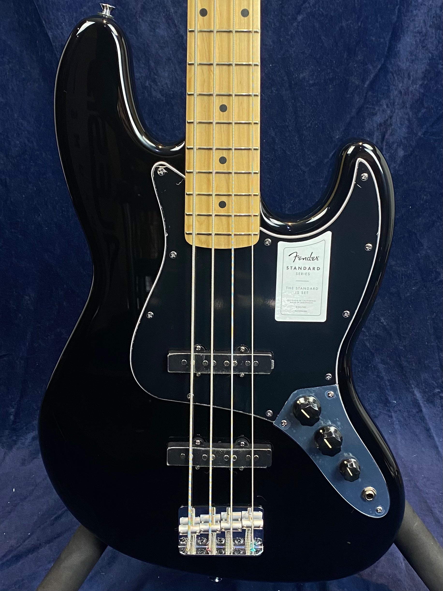Glossy black Fender electric bass with a maple neck, chrome bridge, three control knobs, and a black pickguard against a dark blue background.