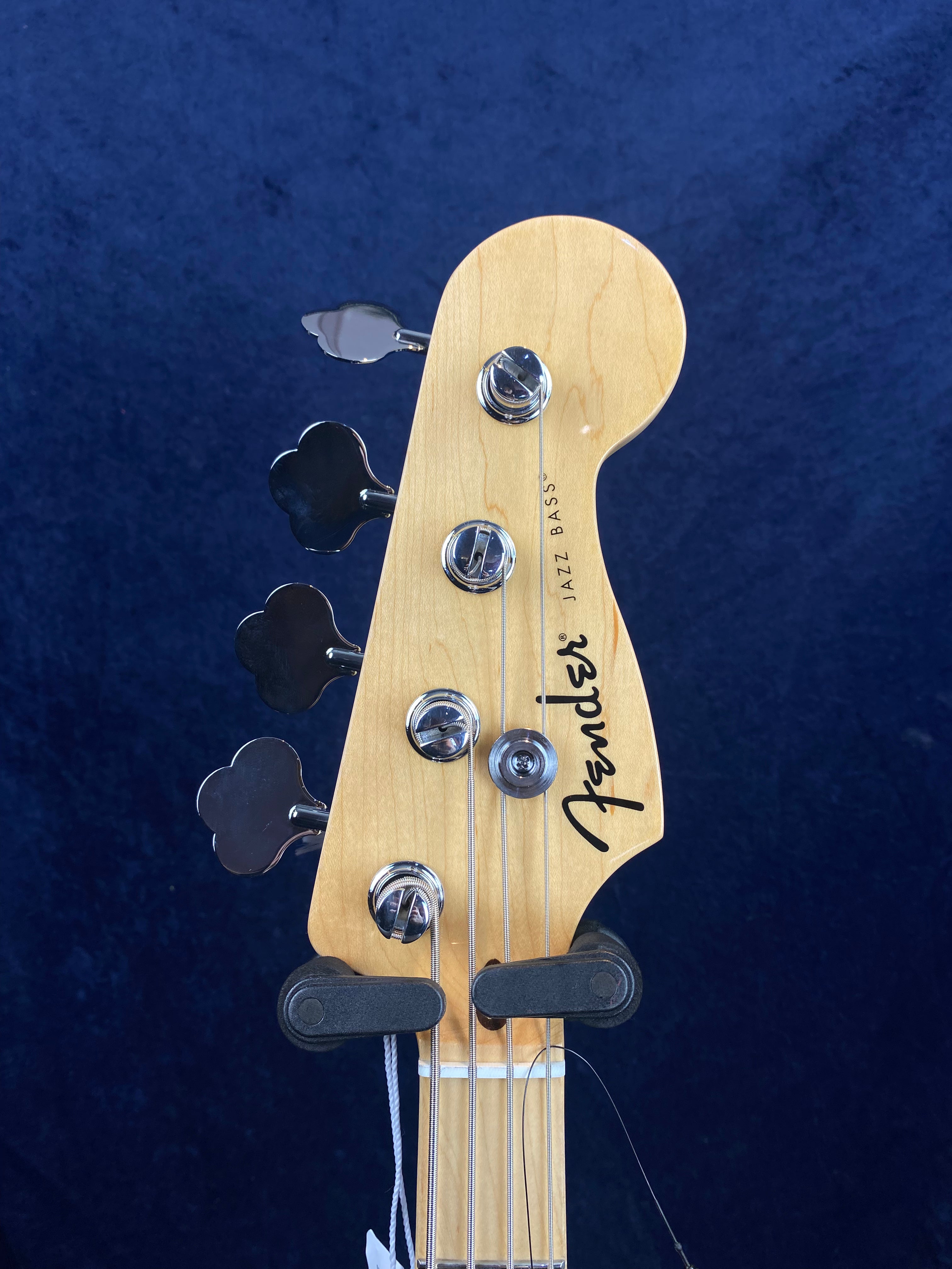 Light wood headstock of a Fender Jazz Bass with four chrome tuning pegs and black logo, shown against a dark blue background.