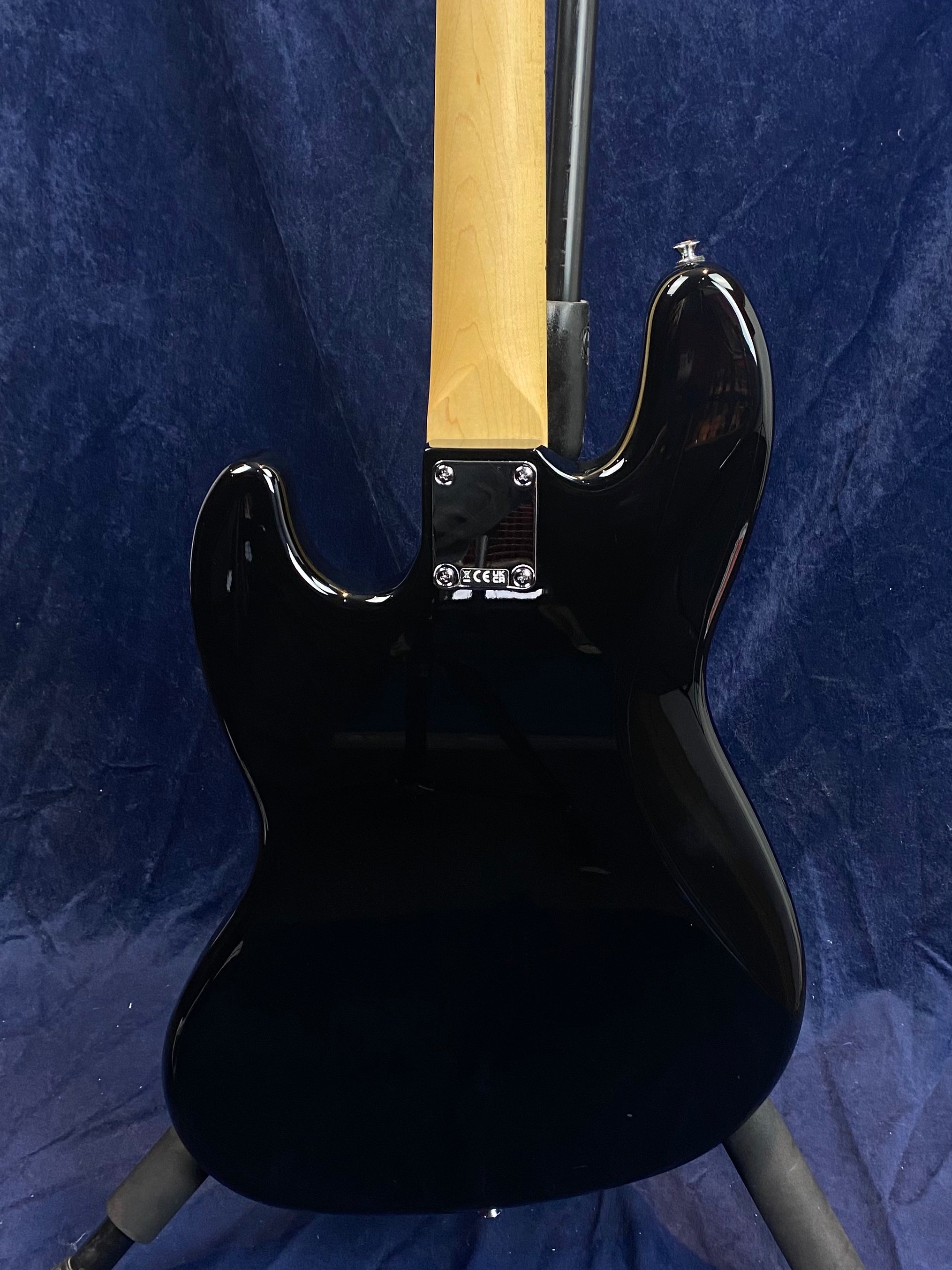 The back of a black electric bass with a natural wood neck, chrome neck plate, and body curves visible against a dark blue background.
