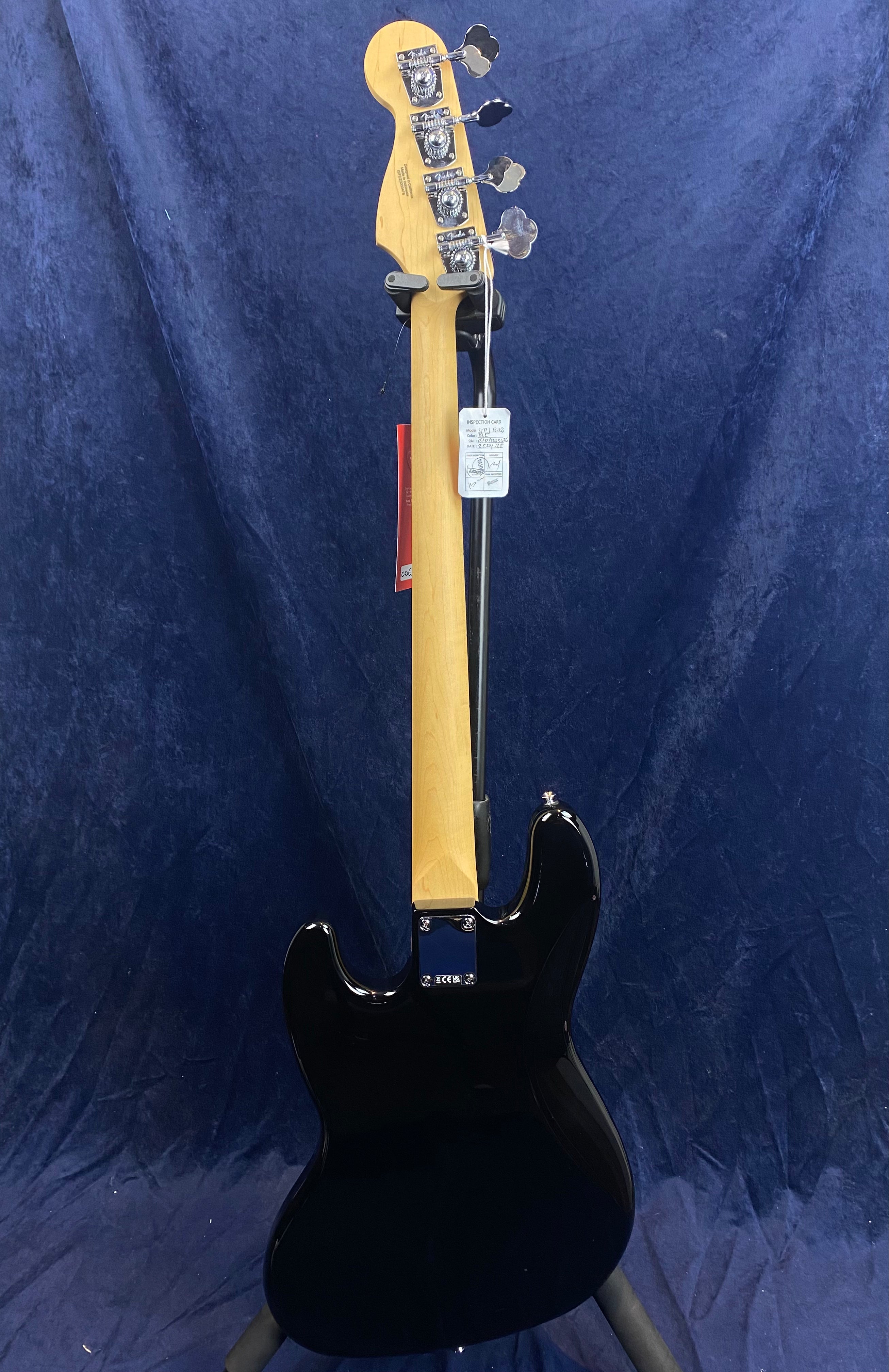 Black electric bass guitar shown from the back, displaying a light wood neck, four tuning pegs, and attached product tags against a dark backdrop.