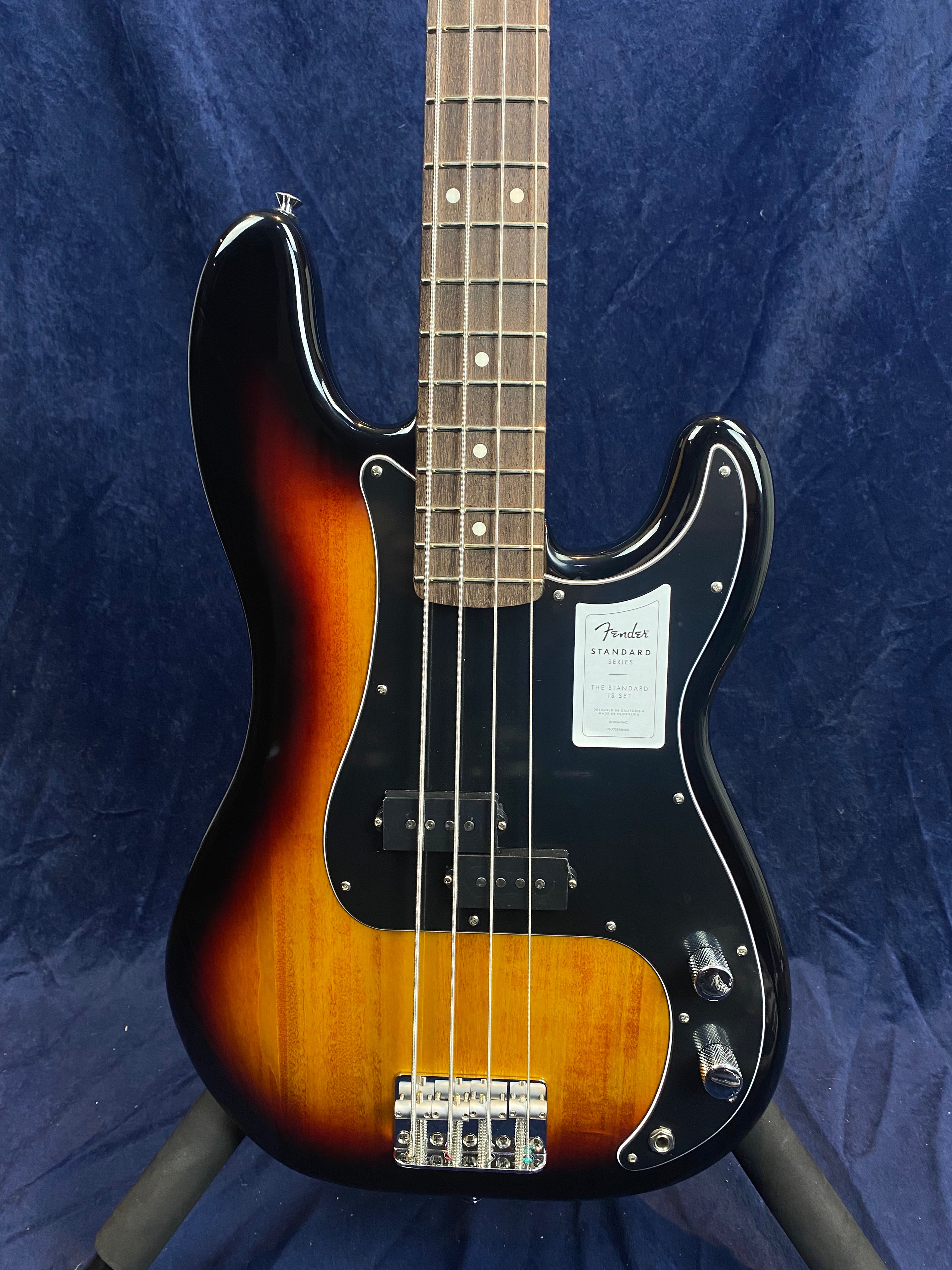 Sunburst Fender electric bass with four strings, black pickguard, two control knobs, and a sticker on the body above the pickups.