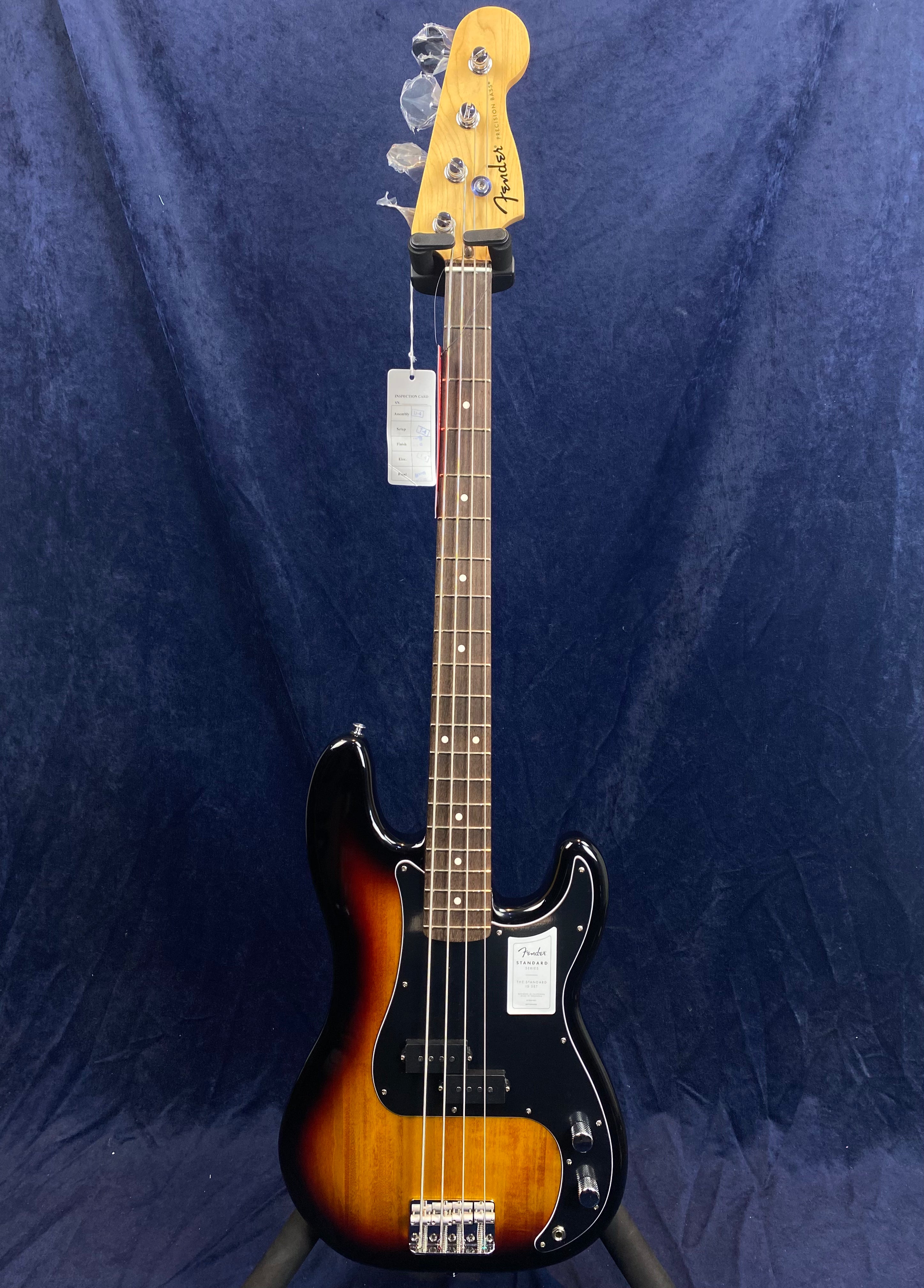 Fender electric bass with a sunburst finish, four strings, black pickguard, and chrome hardware, displayed upright against a dark blue background.