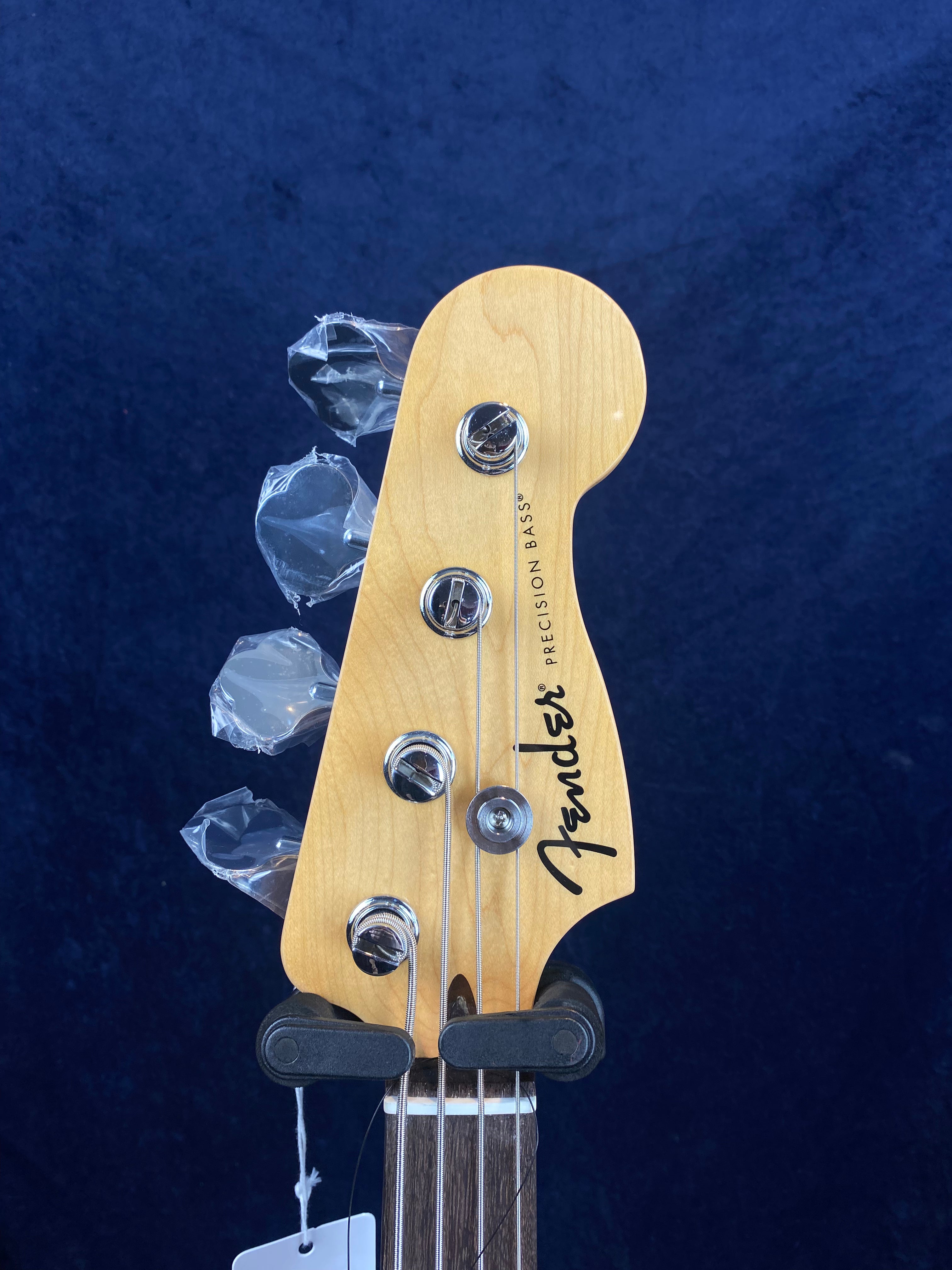 Fender Precision Bass headstock with four tuning pegs, some wrapped in protective plastic, against a dark blue background.