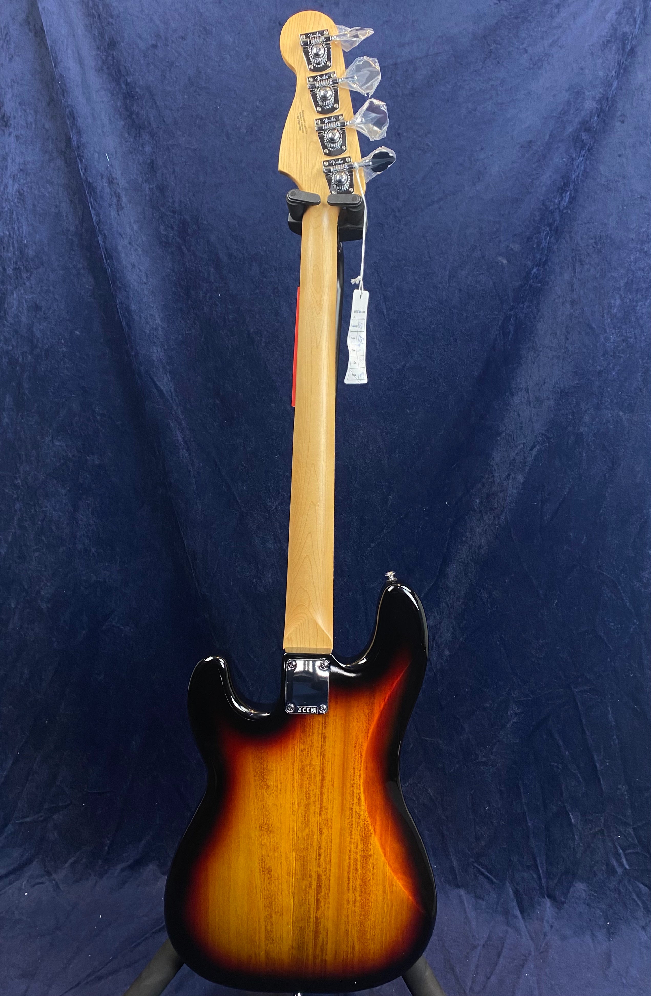 Electric bass seen from the back with a sunburst finish body, light wood neck, and chrome tuning pegs with protective plastic. Tag hangs from the headstock.