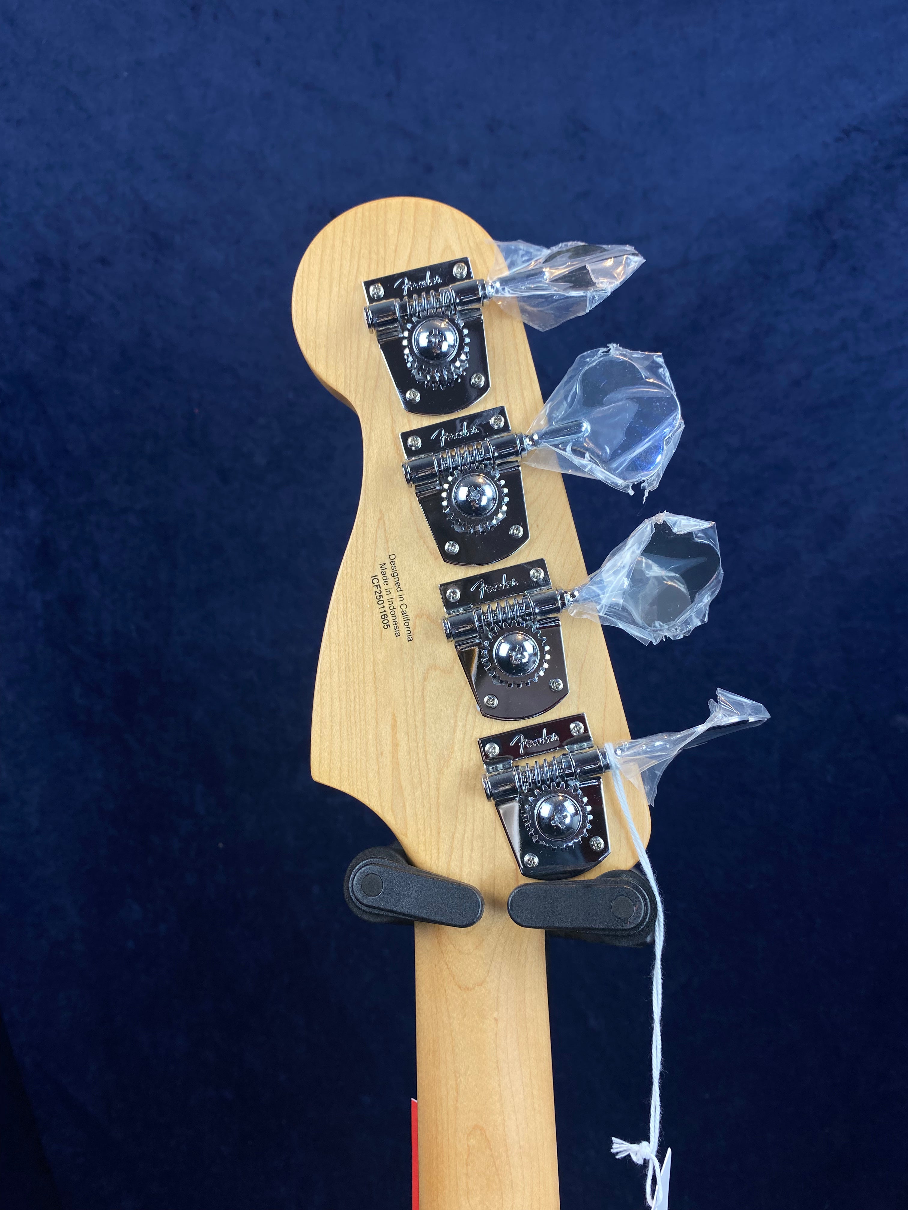 Four chrome tuning pegs with protective plastic wrap on a light wood Fender electric bass headstock against a dark blue background.