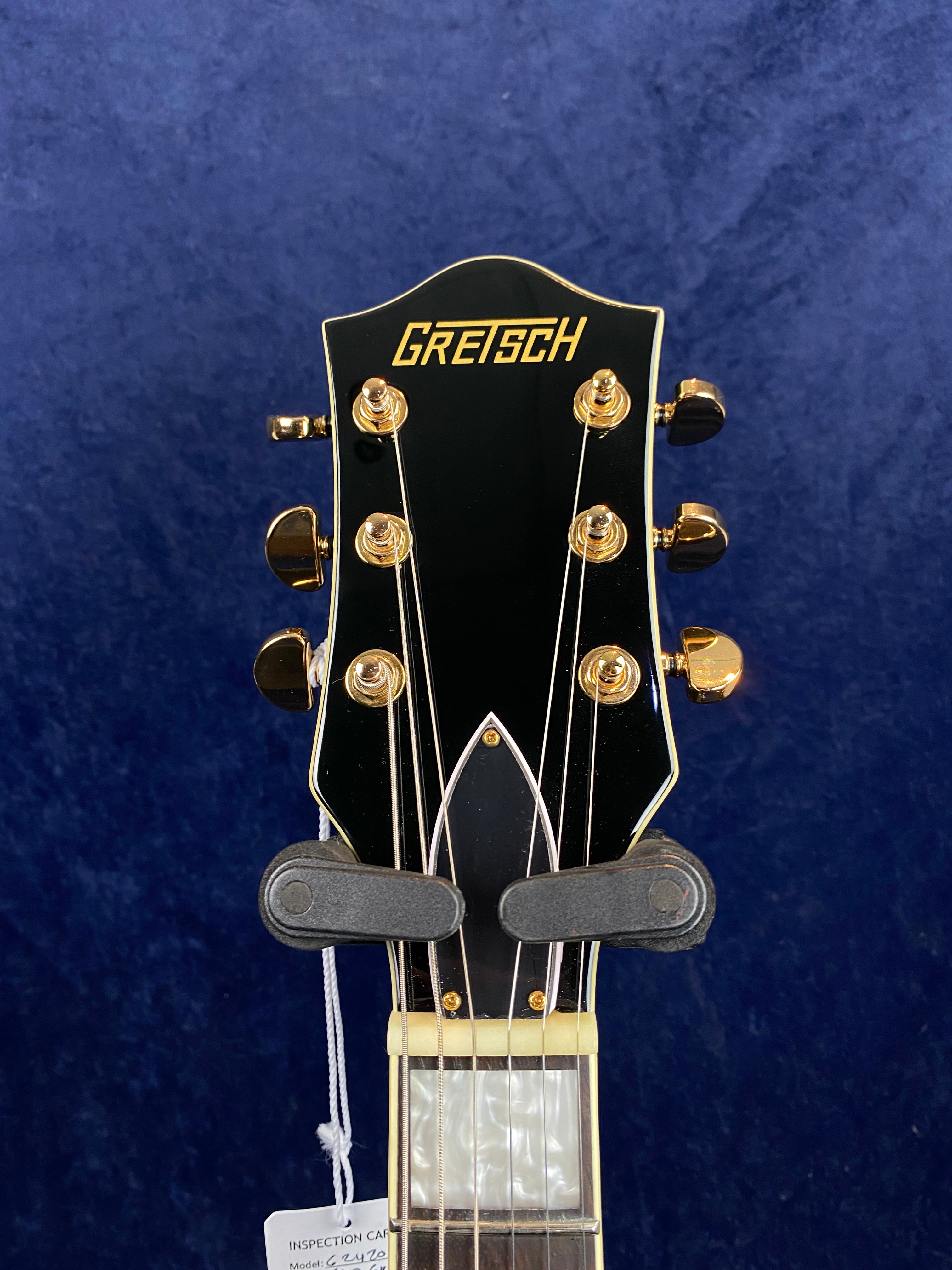 Black Gretsch electric guitar headstock with gold tuning pegs and trim, showing part of the neck and strings, against a dark blue background.