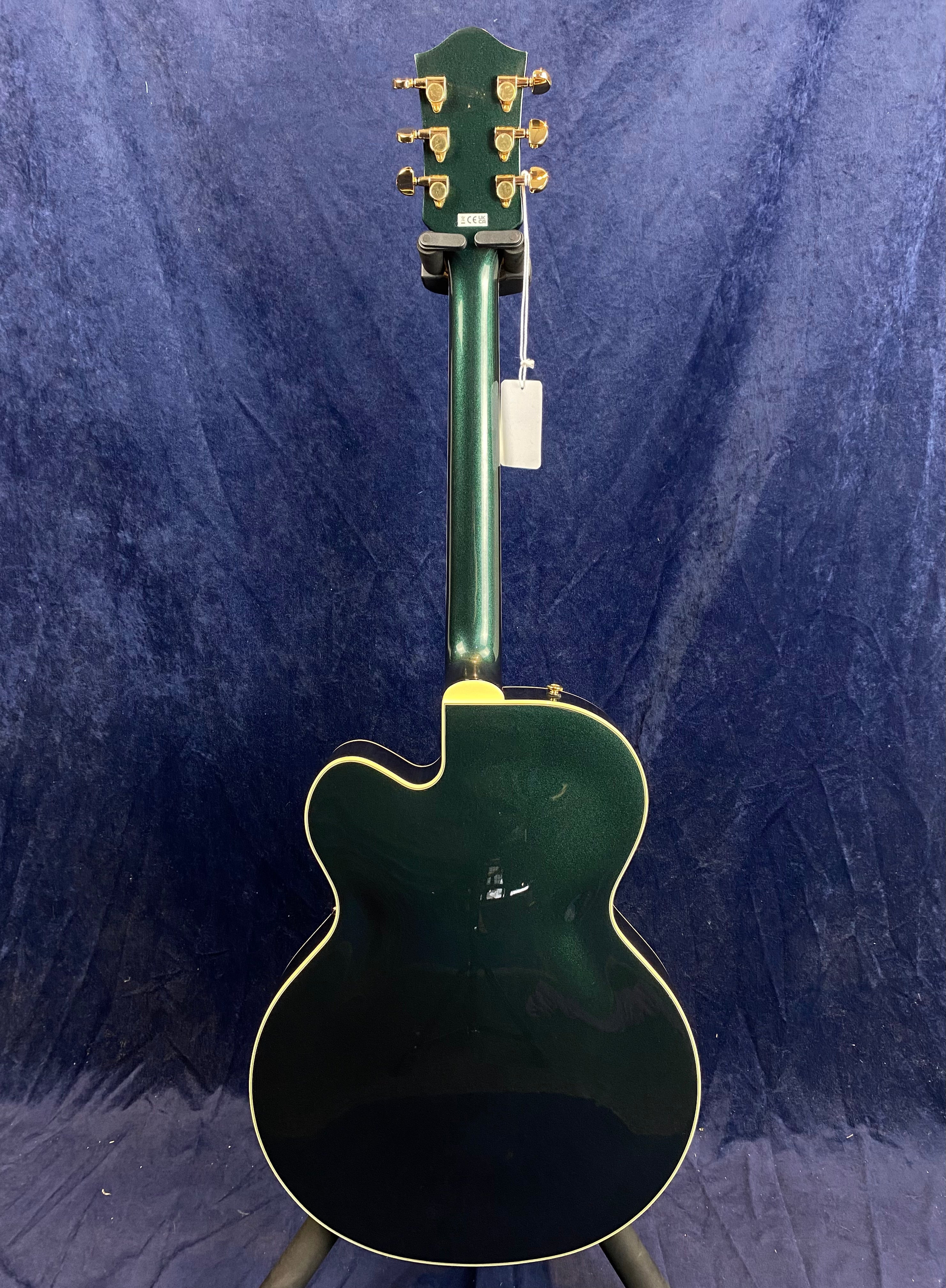 Dark green electric guitar seen from the back, featuring gold tuning pegs and cream binding around the body and neck, set against a blue background.