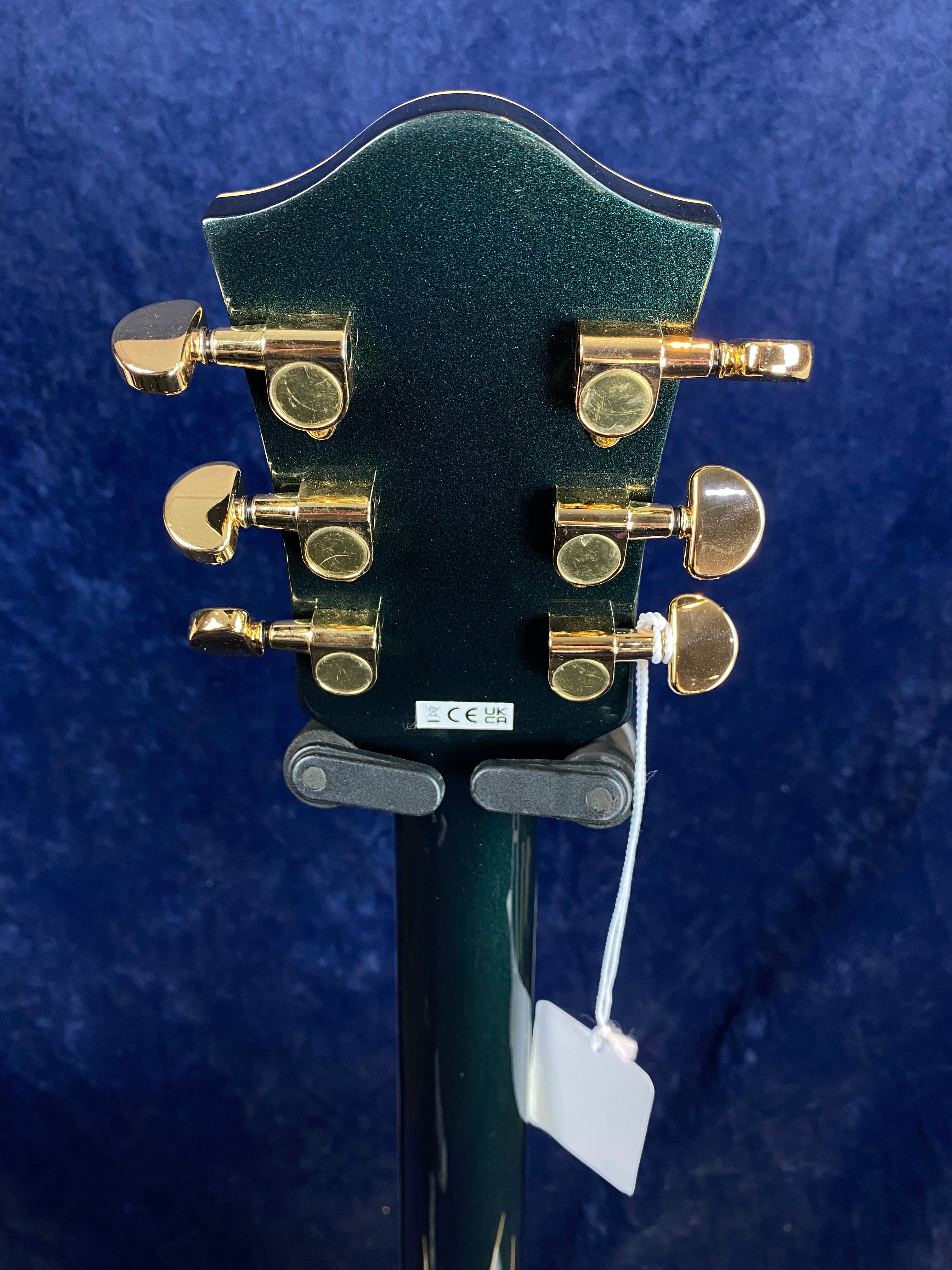 Back of electric guitar headstock with six gold tuning pegs, metallic green finish, and a white tag hanging from the neck.