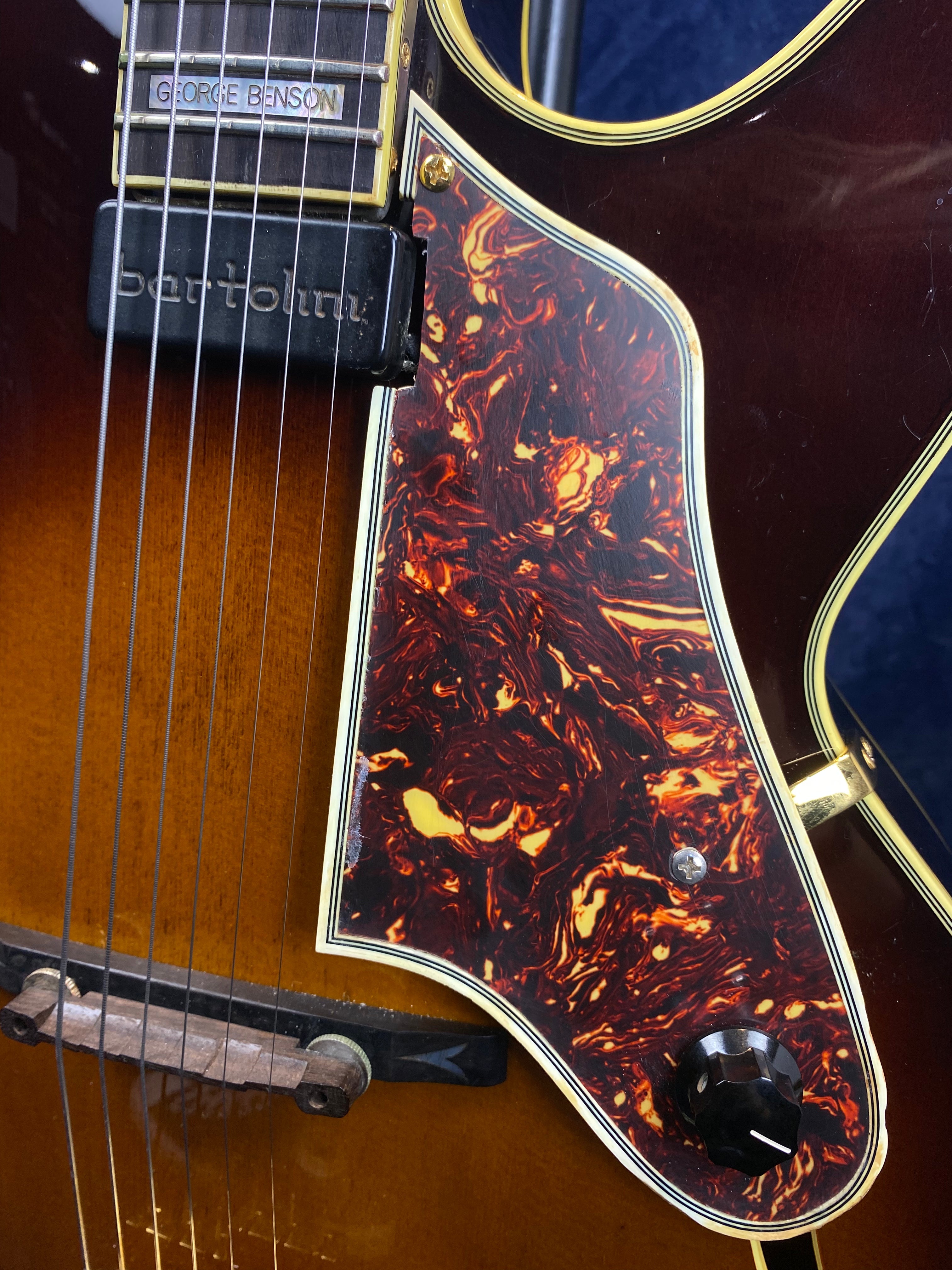 Close-up of an electric guitar with a tortoiseshell-patterned pickguard, George Benson and Bartolini pickups, and a black control knob.