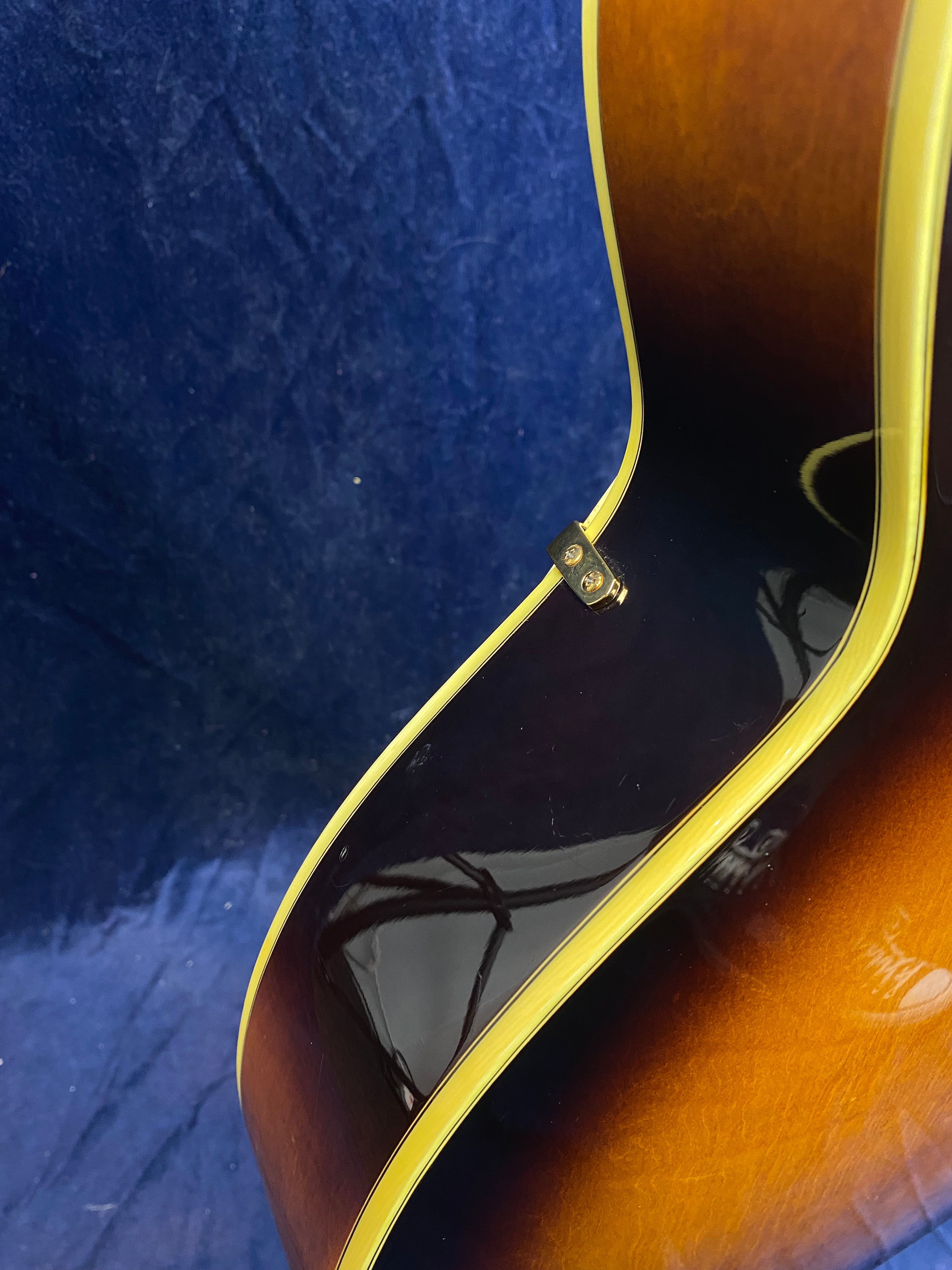 Curved sunburst electric guitar body with glossy finish, cream binding, and a strap button visible against a dark blue background.
