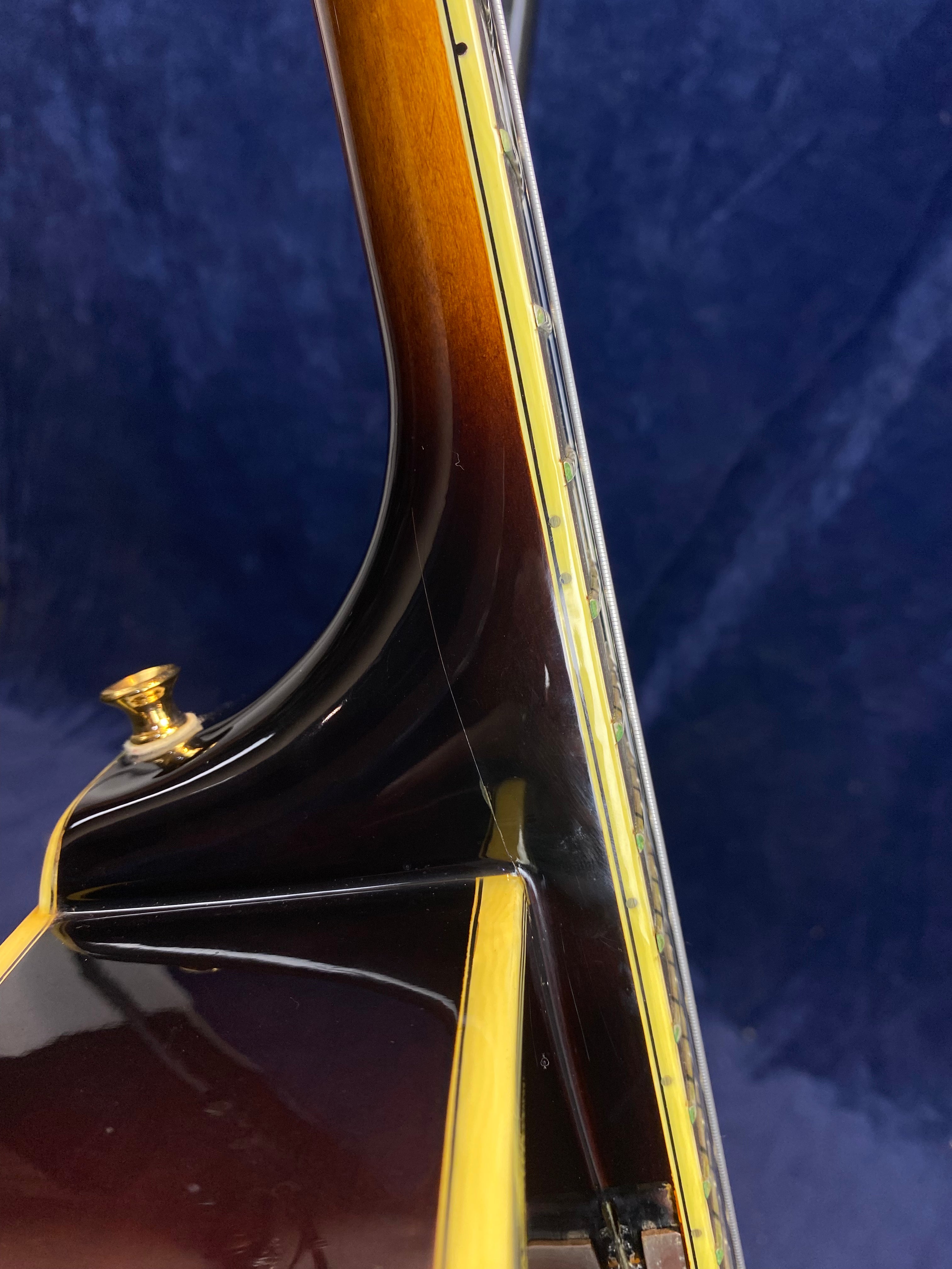 Close-up of an electric guitar’s neck joint with a sunburst finish, cream binding, gold strap button, and visible dot inlays.