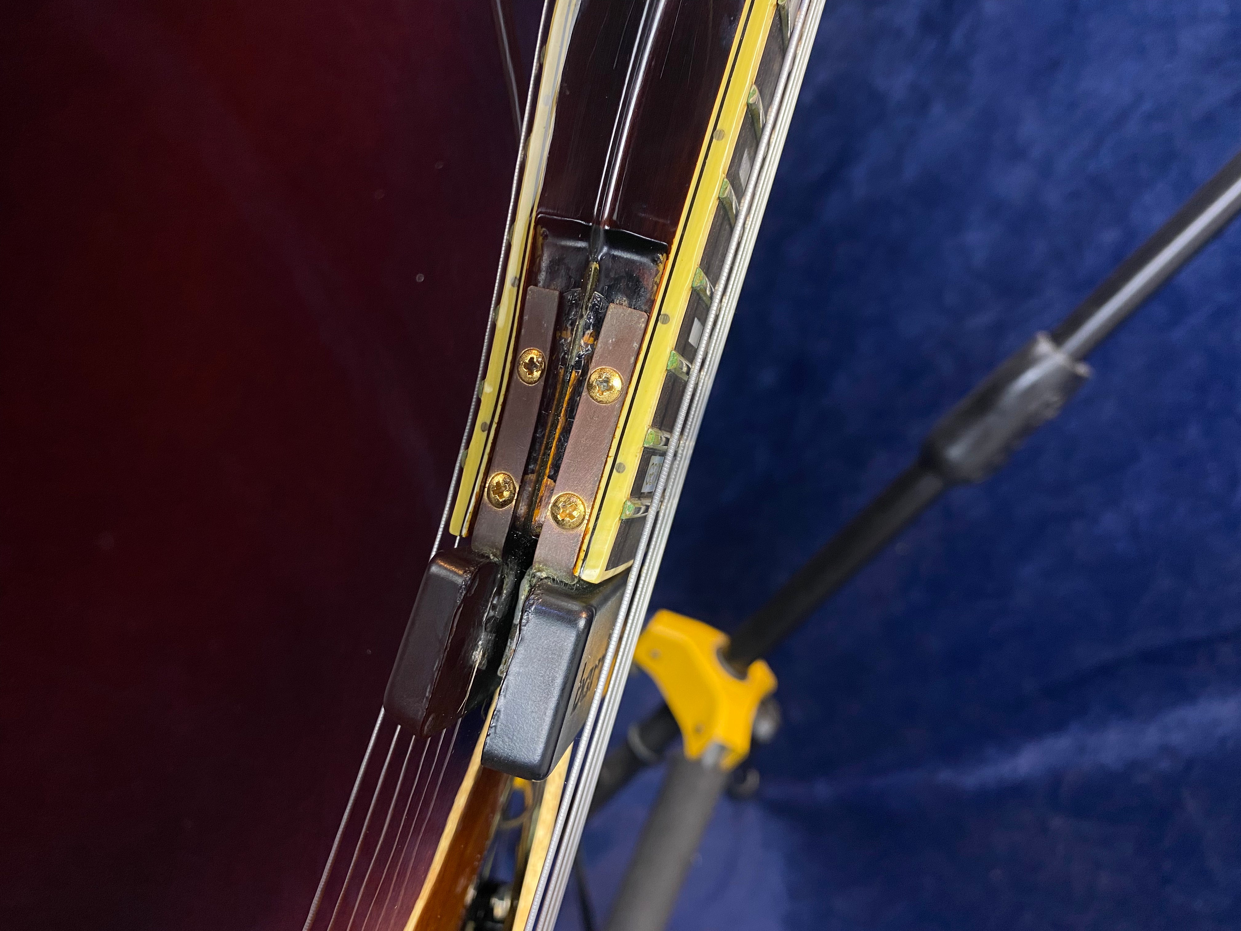 Close-up of an electric guitar’s neck joint showing gold screws, a black hinge mechanism, and yellow binding against a dark background.