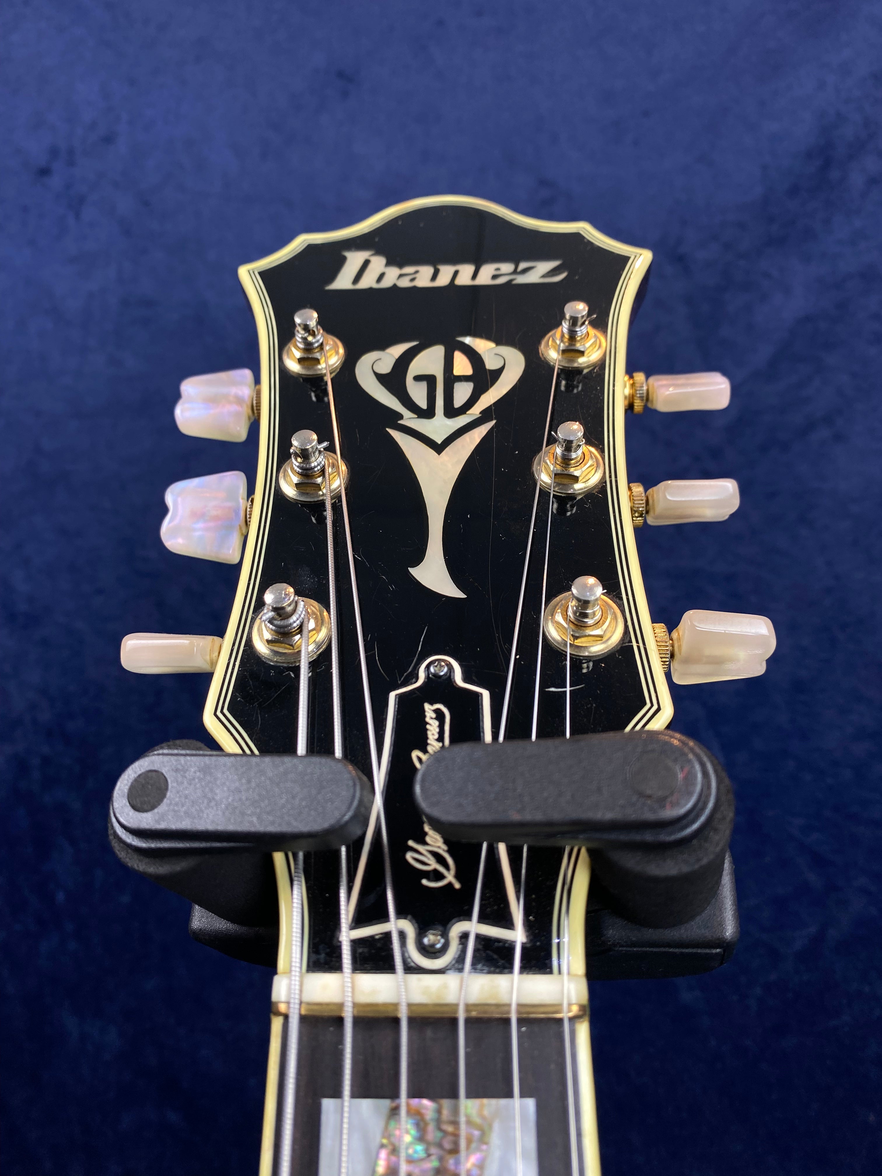 Ibanez electric guitar headstock with gold tuners, pearl tuning pegs, GB inlay, and signature truss rod cover on a dark background.