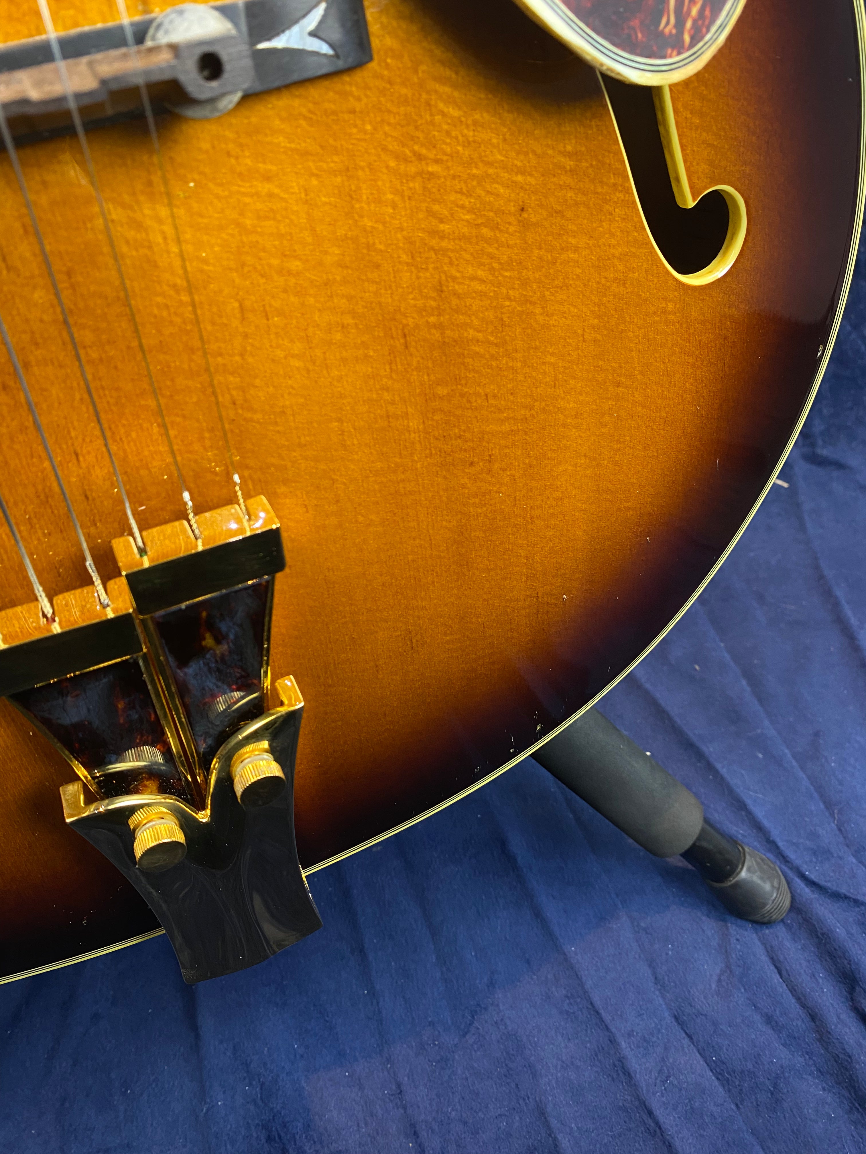 Sunburst finish electric guitar body with gold hardware, visible f-hole, tortoiseshell-style tailpiece, and strings resting on a blue fabric surface.