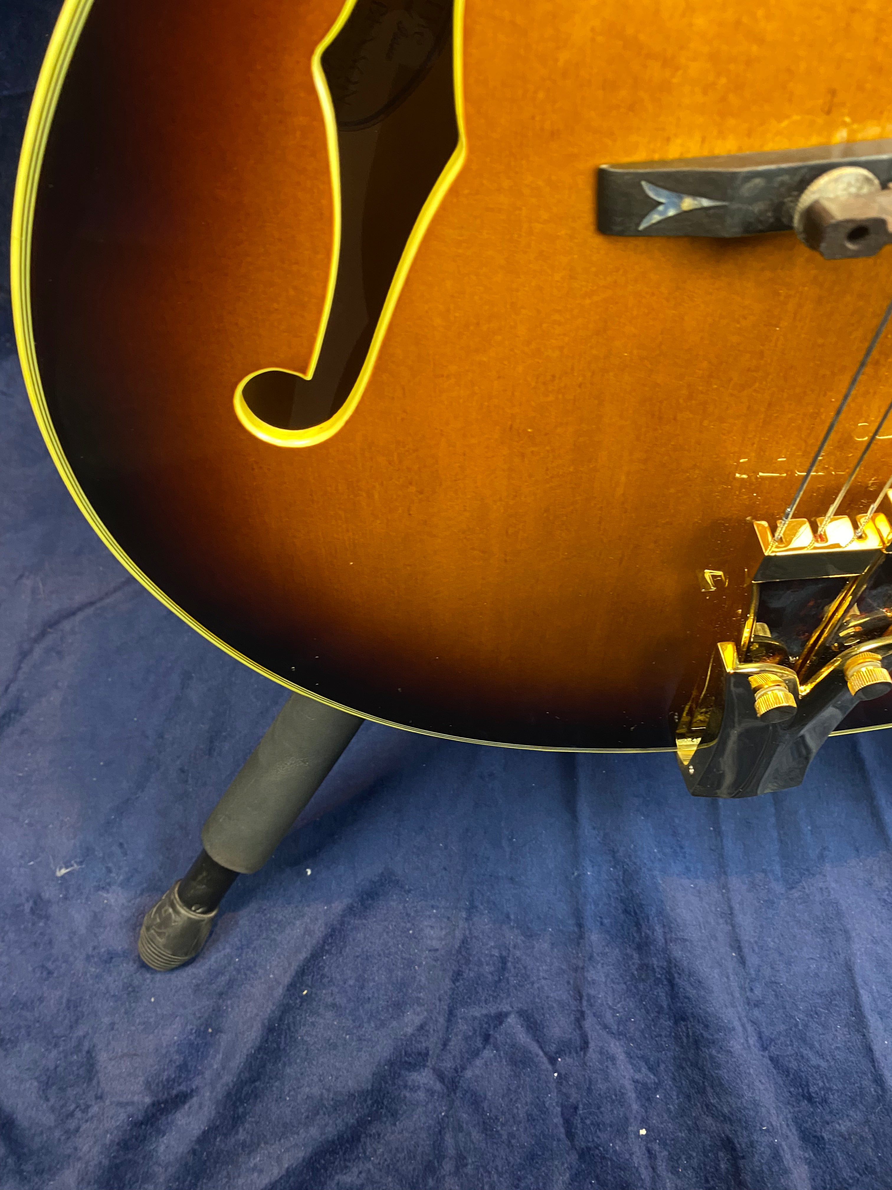 Curved f-hole, sunburst finish, and gold hardware are visible on the body of a hollow electric guitar resting on a stand over blue fabric.