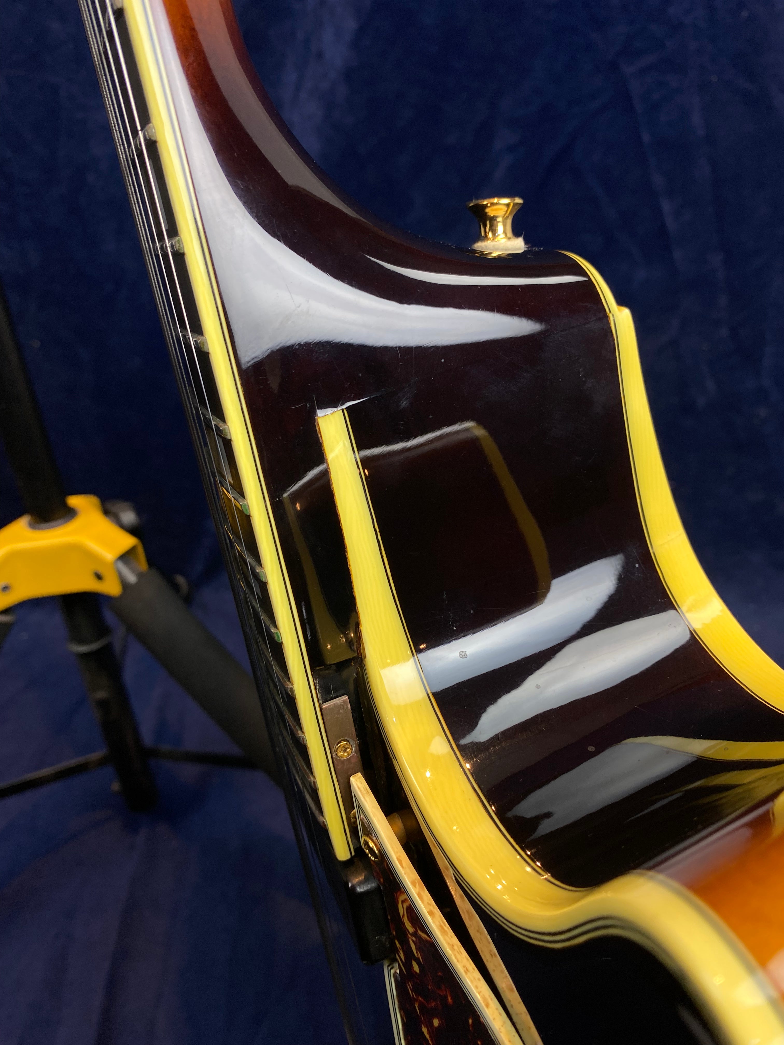 Close-up of an electric guitar’s neck joint, showing glossy dark wood, yellow binding, a gold strap button, and a guitar stand base in the background.
