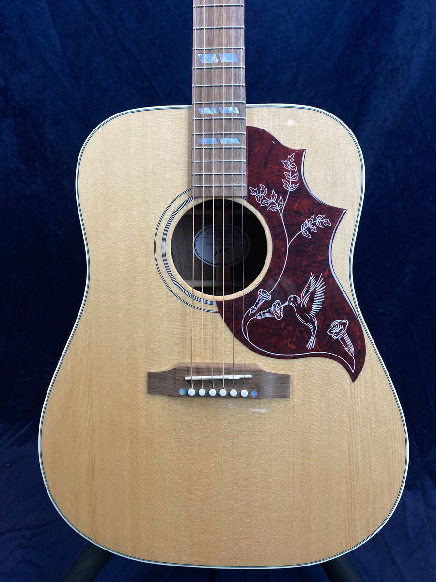 Natural wood acoustic guitar with a decorative red pickguard featuring a white hummingbird and flowers design.