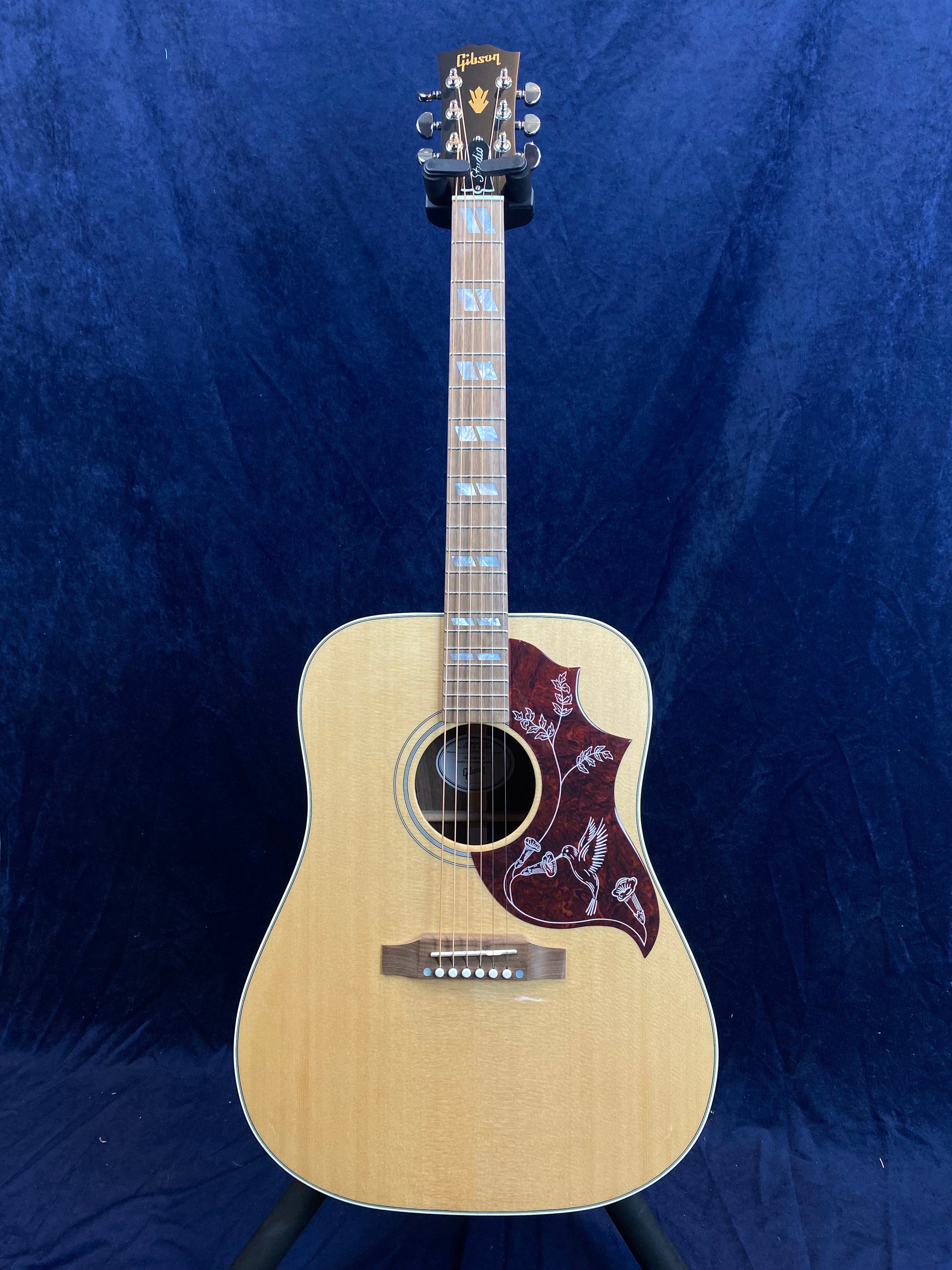 Natural wood Gibson acoustic guitar with decorative hummingbird pickguard, mother-of-pearl inlays on the neck, and gold Gibson logo on the headstock.