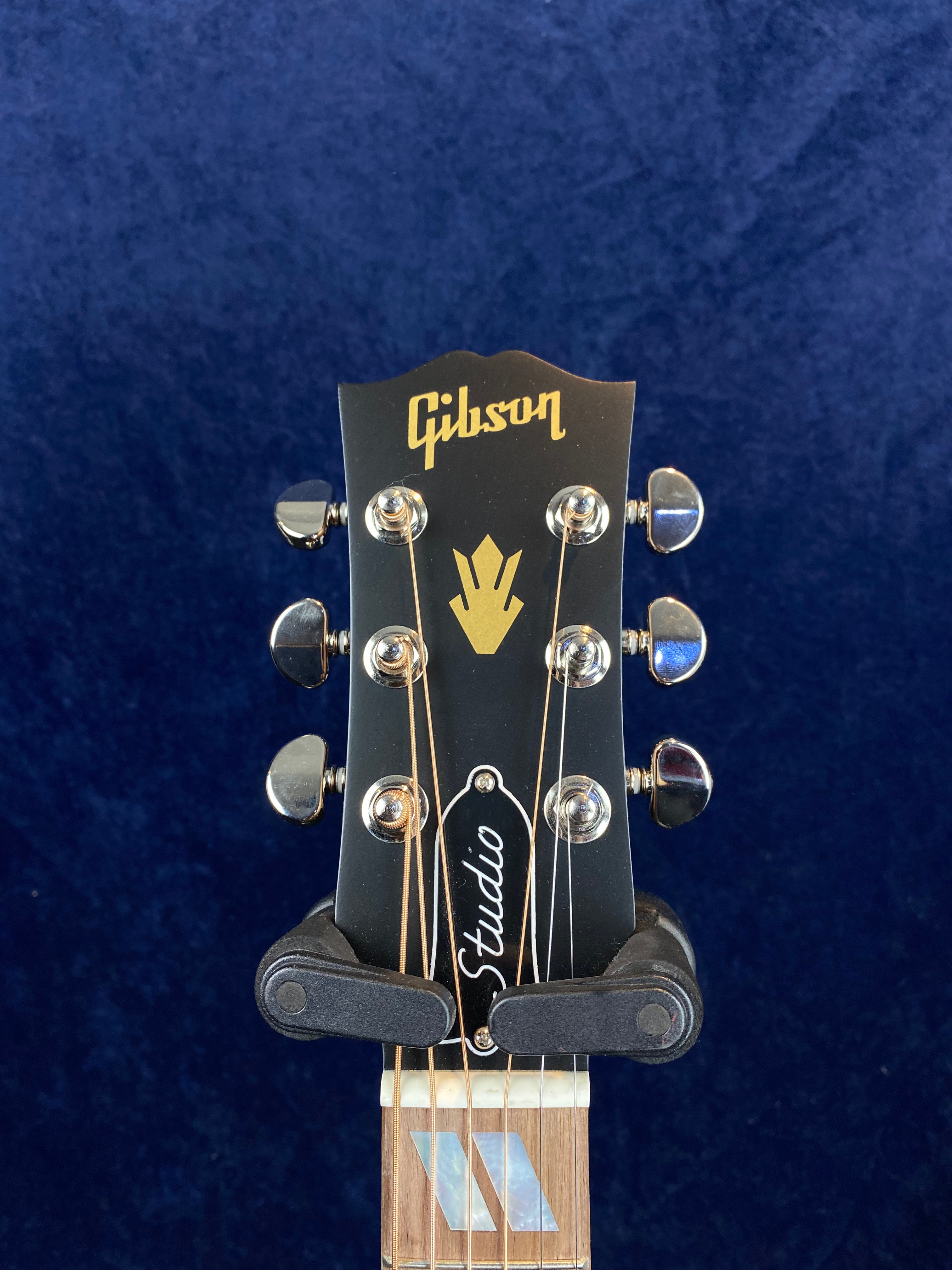 Black Gibson acoustic guitar headstock with gold logo, silver tuning pegs, Studio label, and geometric inlay on the fretboard.