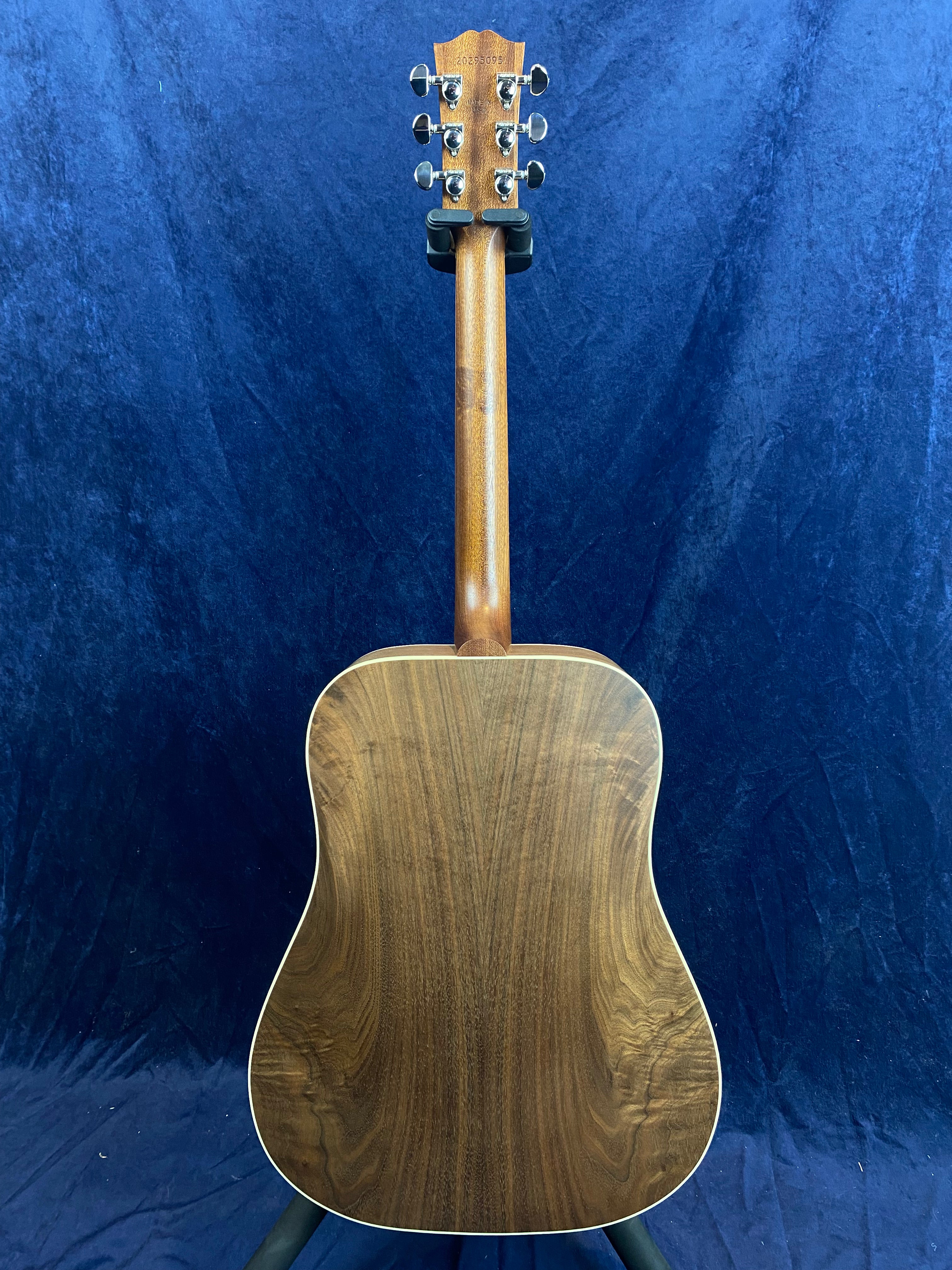Acoustic guitar with a smooth, dark wood grain back and neck, displayed upright against a deep blue background. Silver tuning pegs visible on headstock.