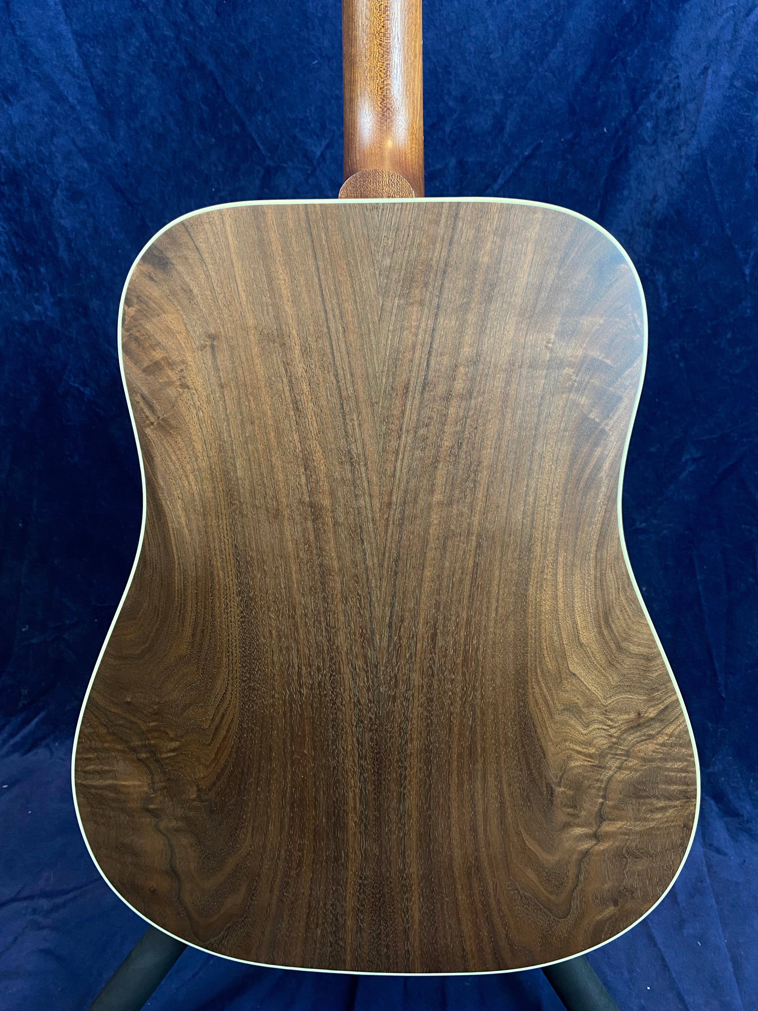 Acoustic guitar back with natural dark wood grain, slightly curved shape, cream binding along the edges, and mahogany neck visible at the top.