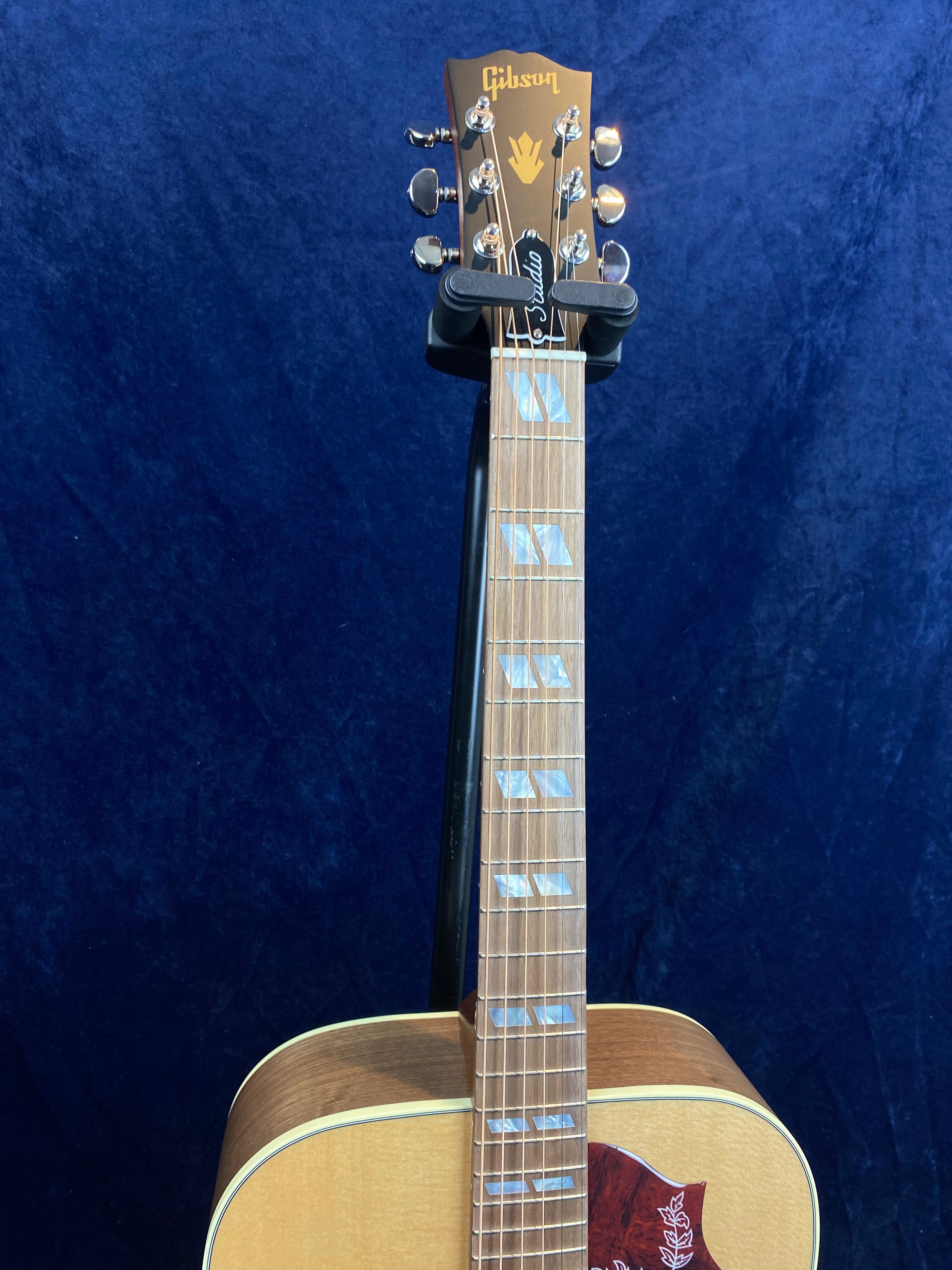 Gibson acoustic guitar with a light wood body, pearl inlays on the fretboard, and a dark red pickguard featuring a decorative white design.