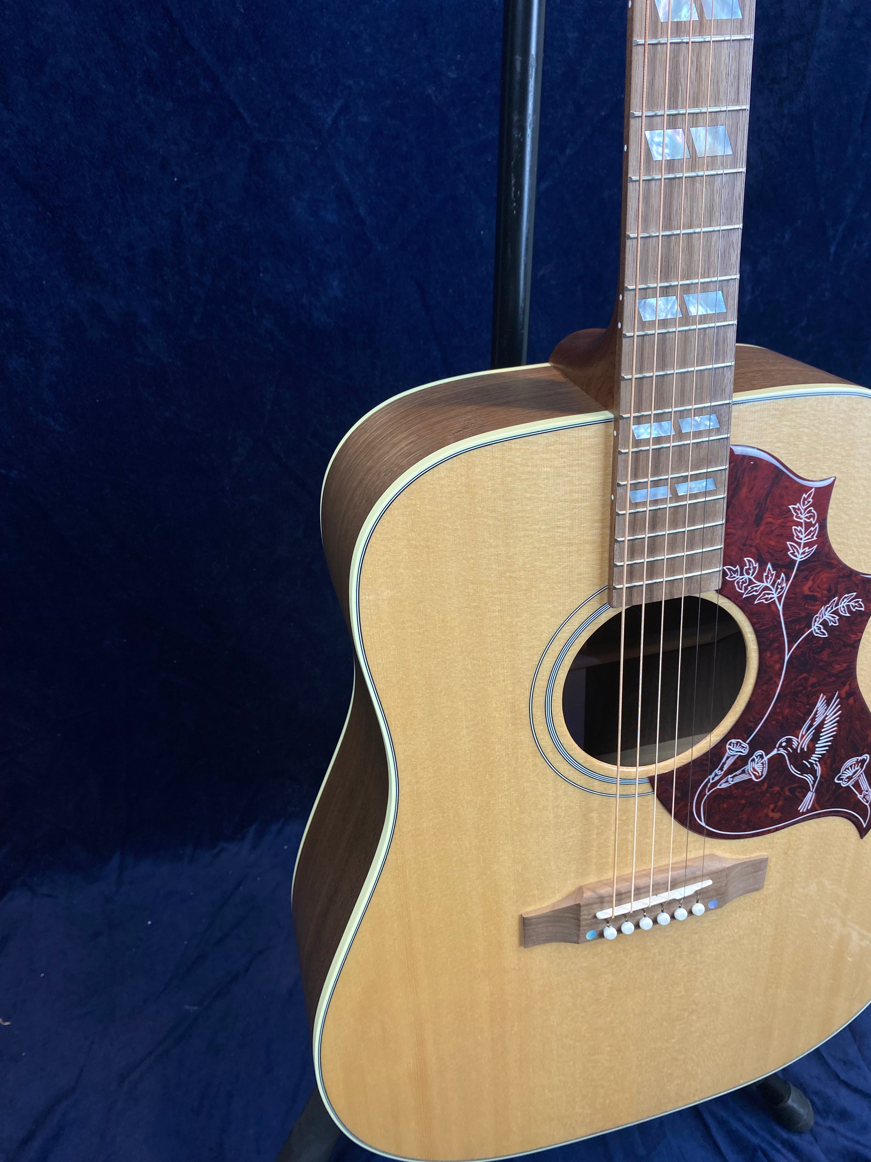 Light wood acoustic guitar with a hummingbird and floral design on the pickguard, white binding, and pearl inlays on the fretboard.