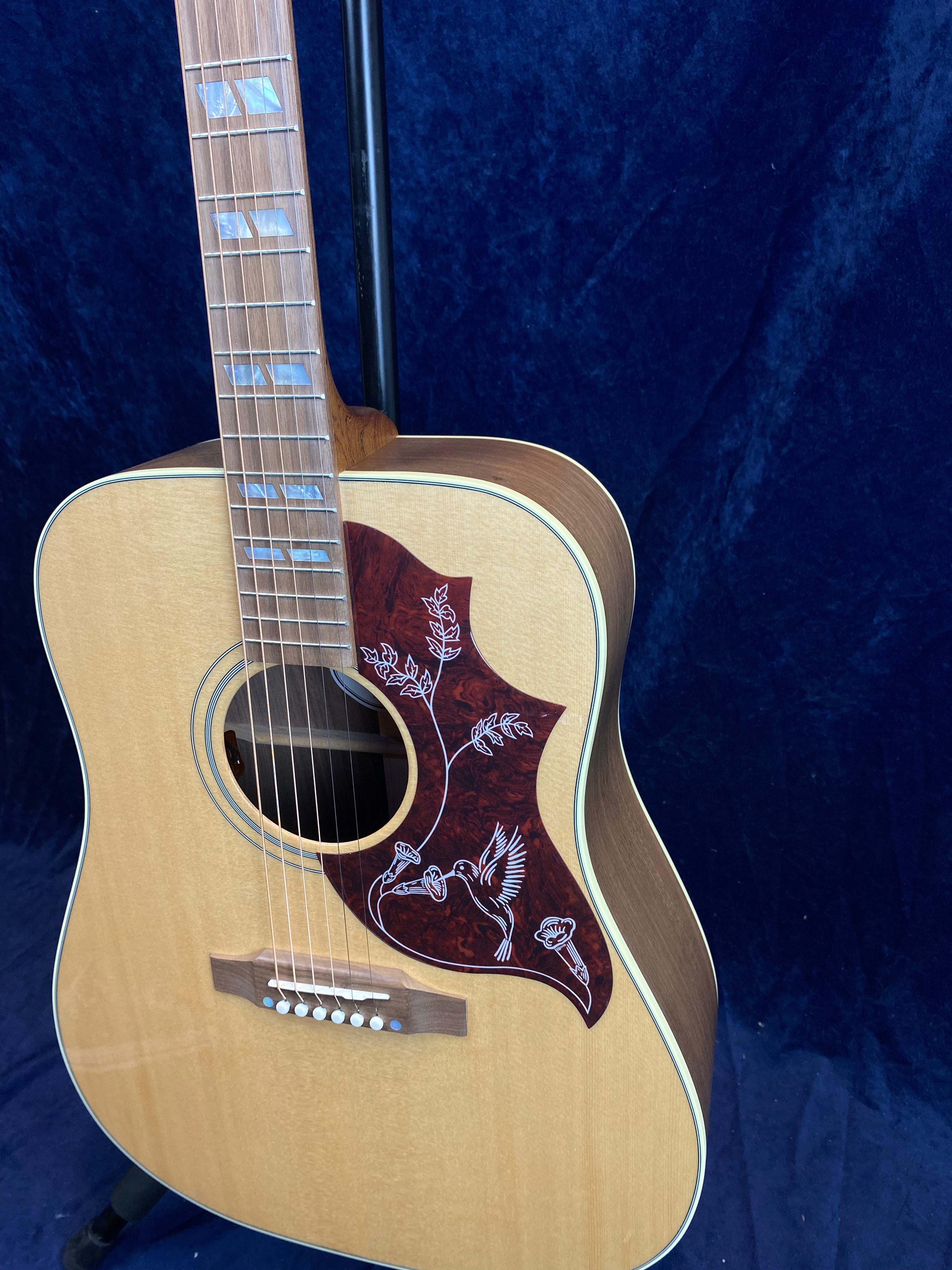 Natural wood acoustic guitar with a reddish pickguard featuring a floral design and a bird, and rectangular mother-of-pearl inlays on the fretboard.