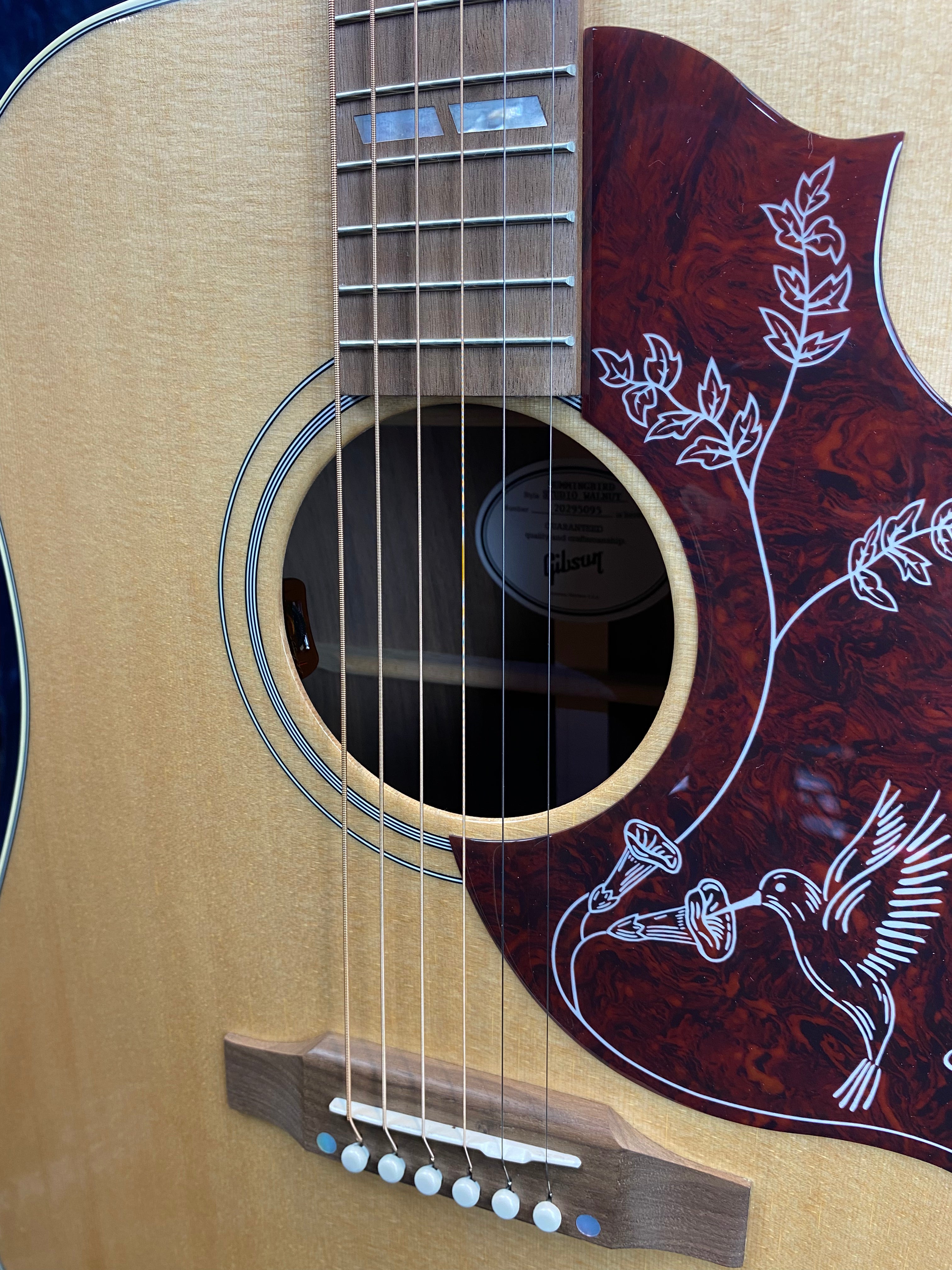Light wood acoustic guitar with a red pickguard featuring a white hummingbird and flowers design. Six steel strings and mother-of-pearl inlays on the fretboard.